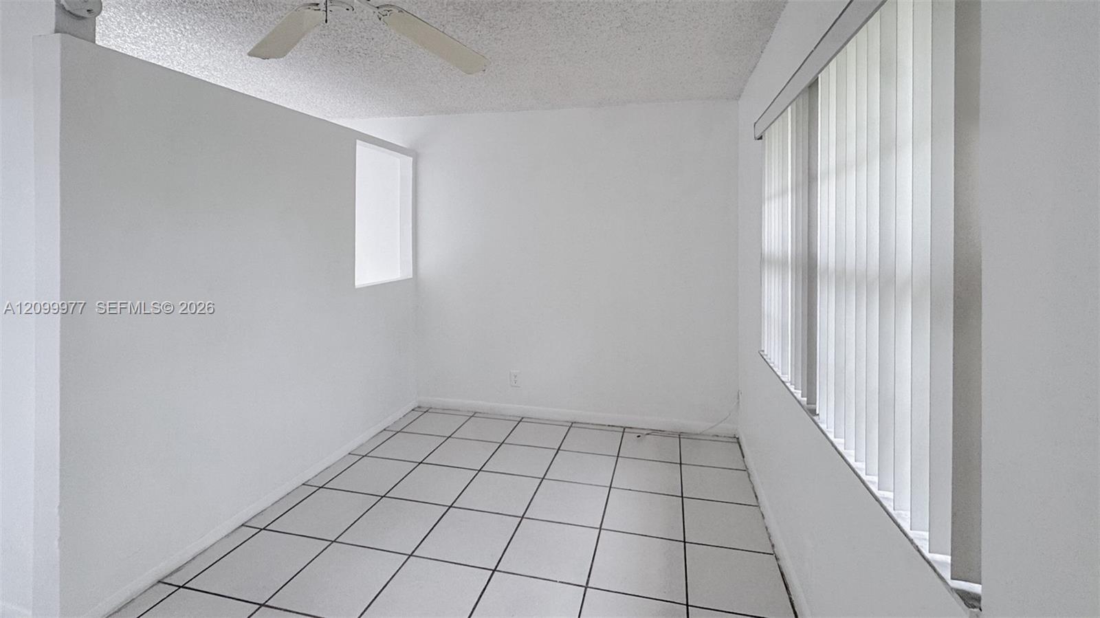Empty tiled room with a ceiling fan and vertical blinds over a large window.