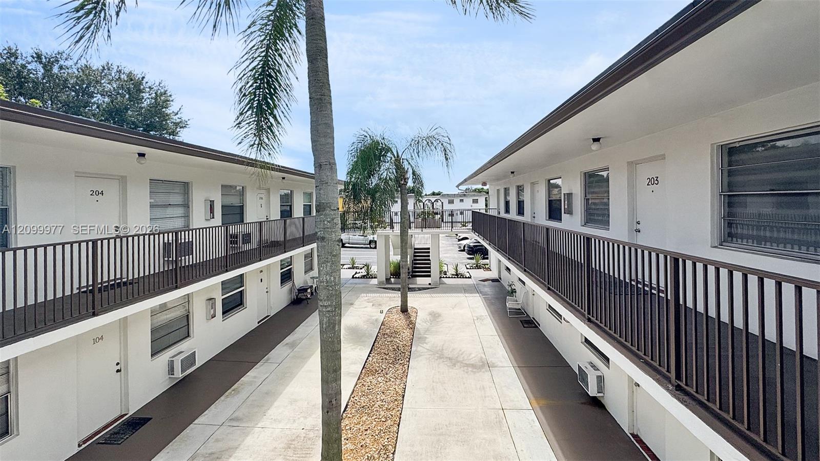 Apartment courtyard between white two-story buildings, with palm trees, exterior walkways, and a central stairway.