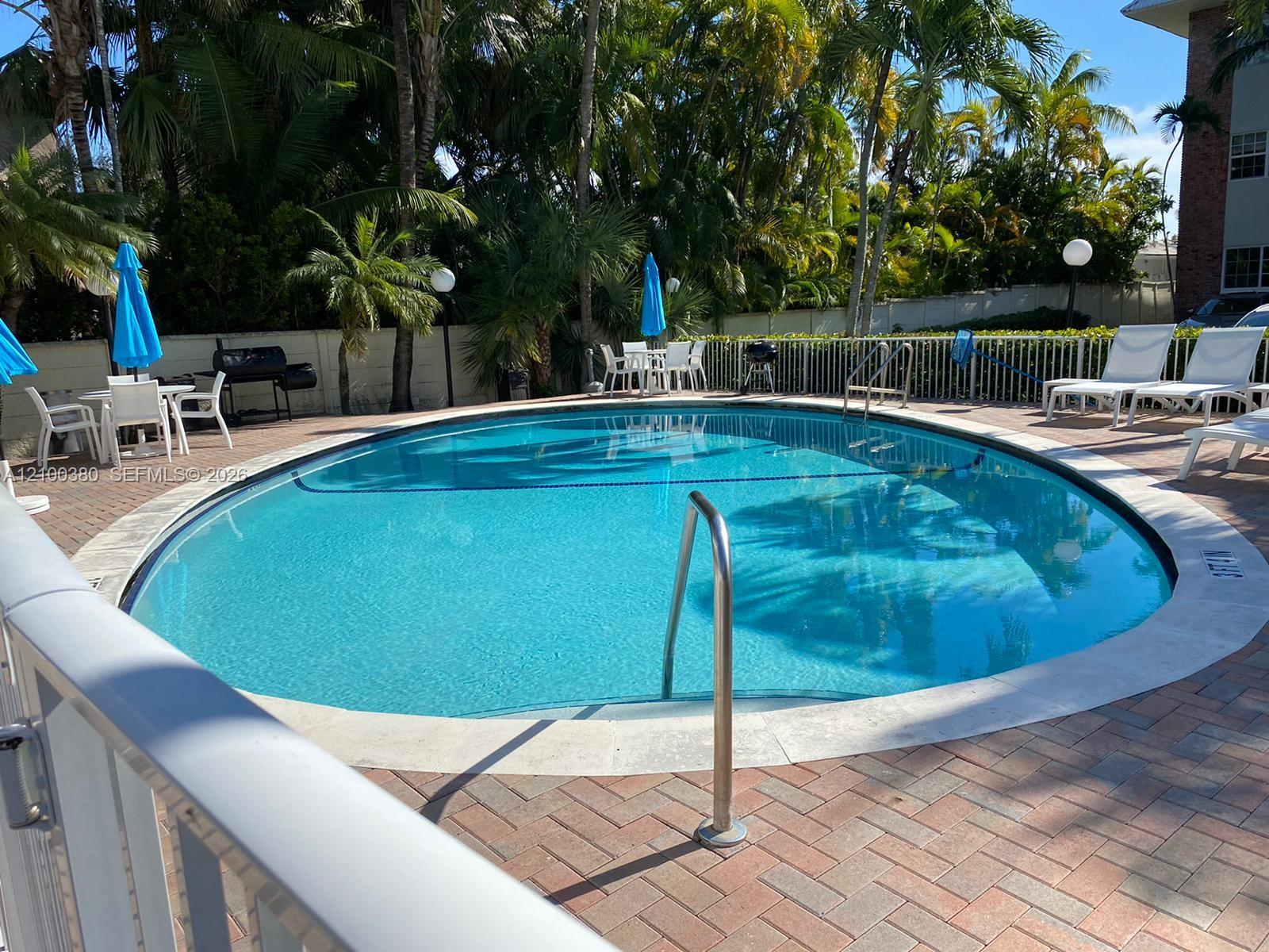 Outdoor pool surrounded by palm trees, lounge chairs, and blue umbrellas.