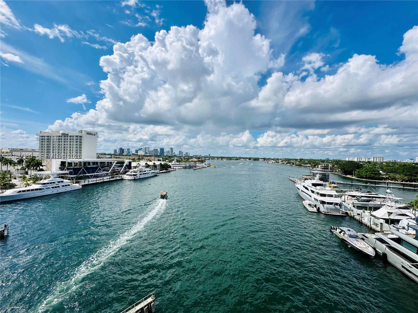 Waterway view with moored yachts, a boat wake, and a distant city skyline.