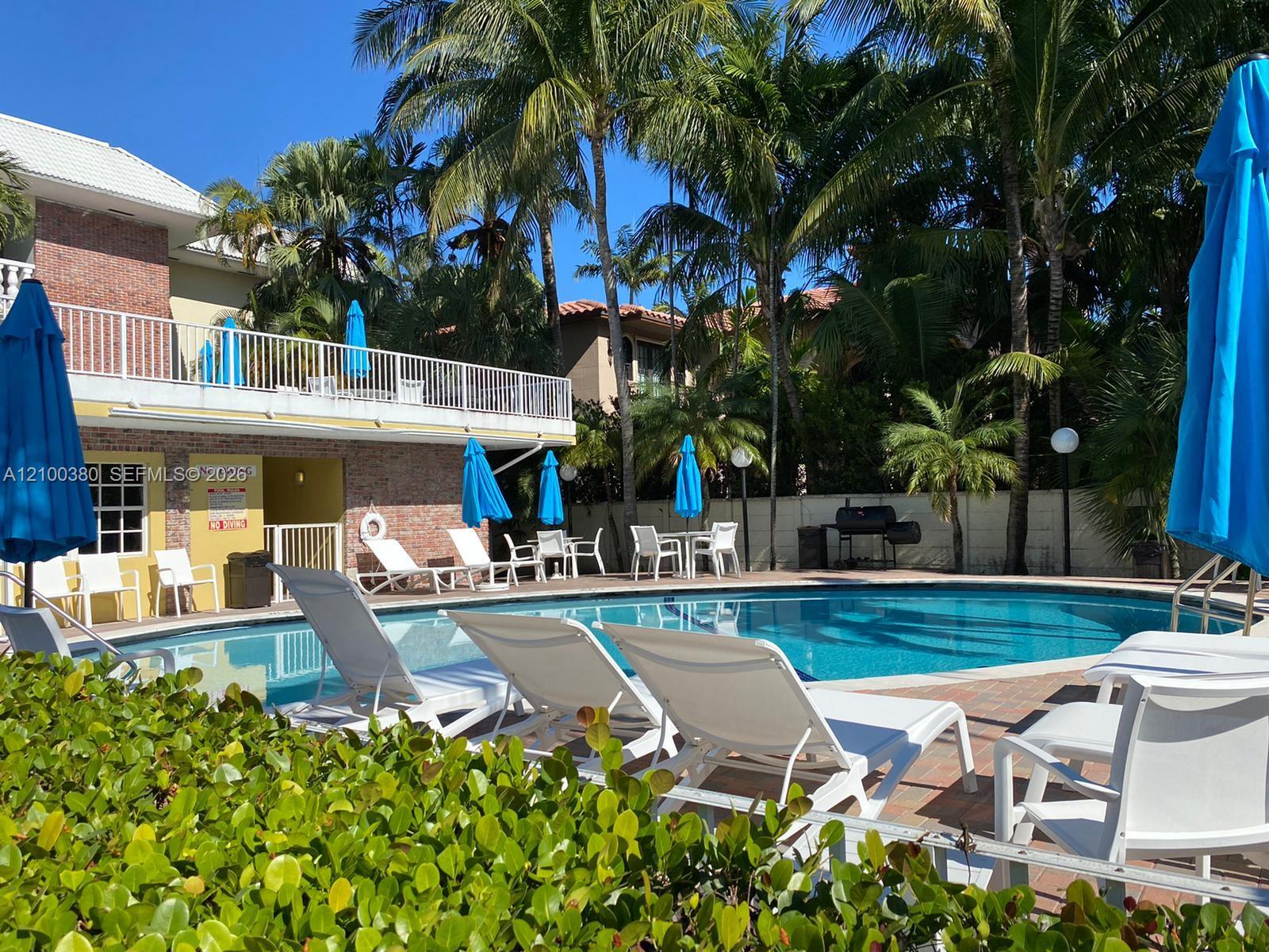 Condominium pool with white lounge chairs, blue umbrellas, and surrounding palm trees.