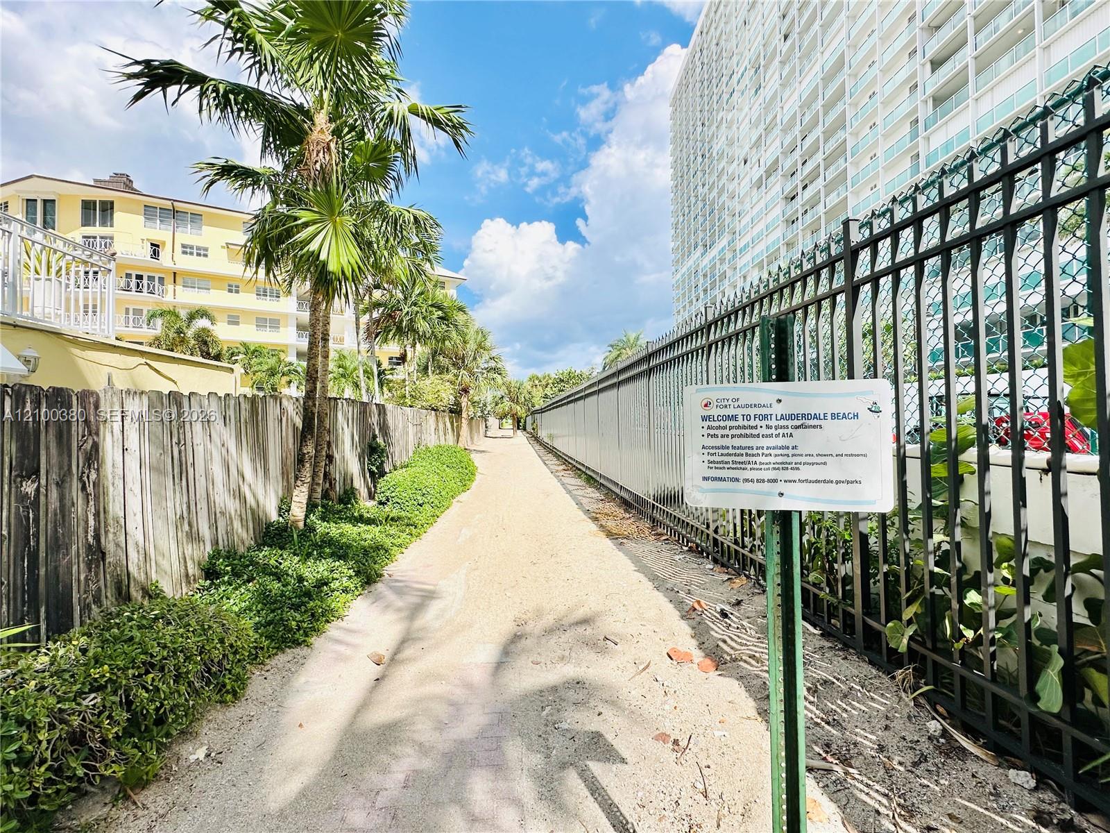 Walkway beside Fort Lauderdale condominiums, lined with palms and marked by a Fort Lauderdale Beach sign.