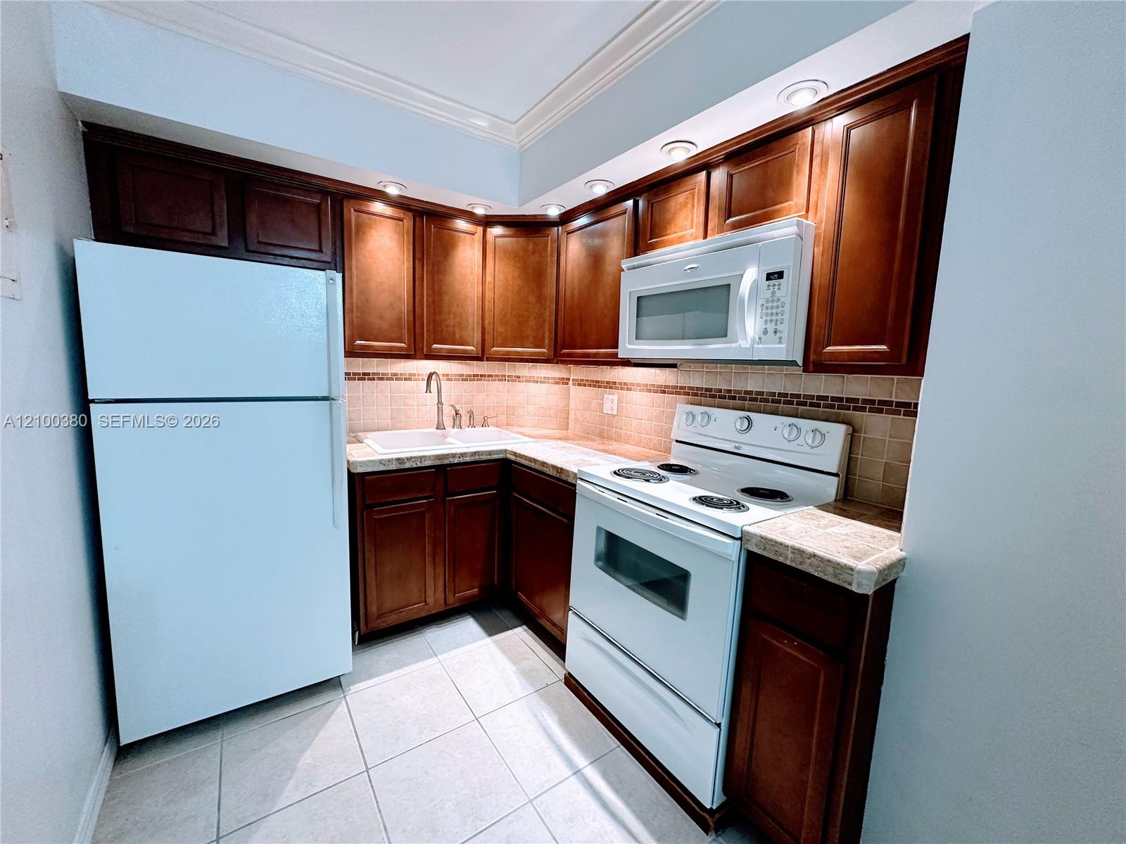 Kitchen with dark wood cabinets, tiled counters, white appliances, and a corner sink.