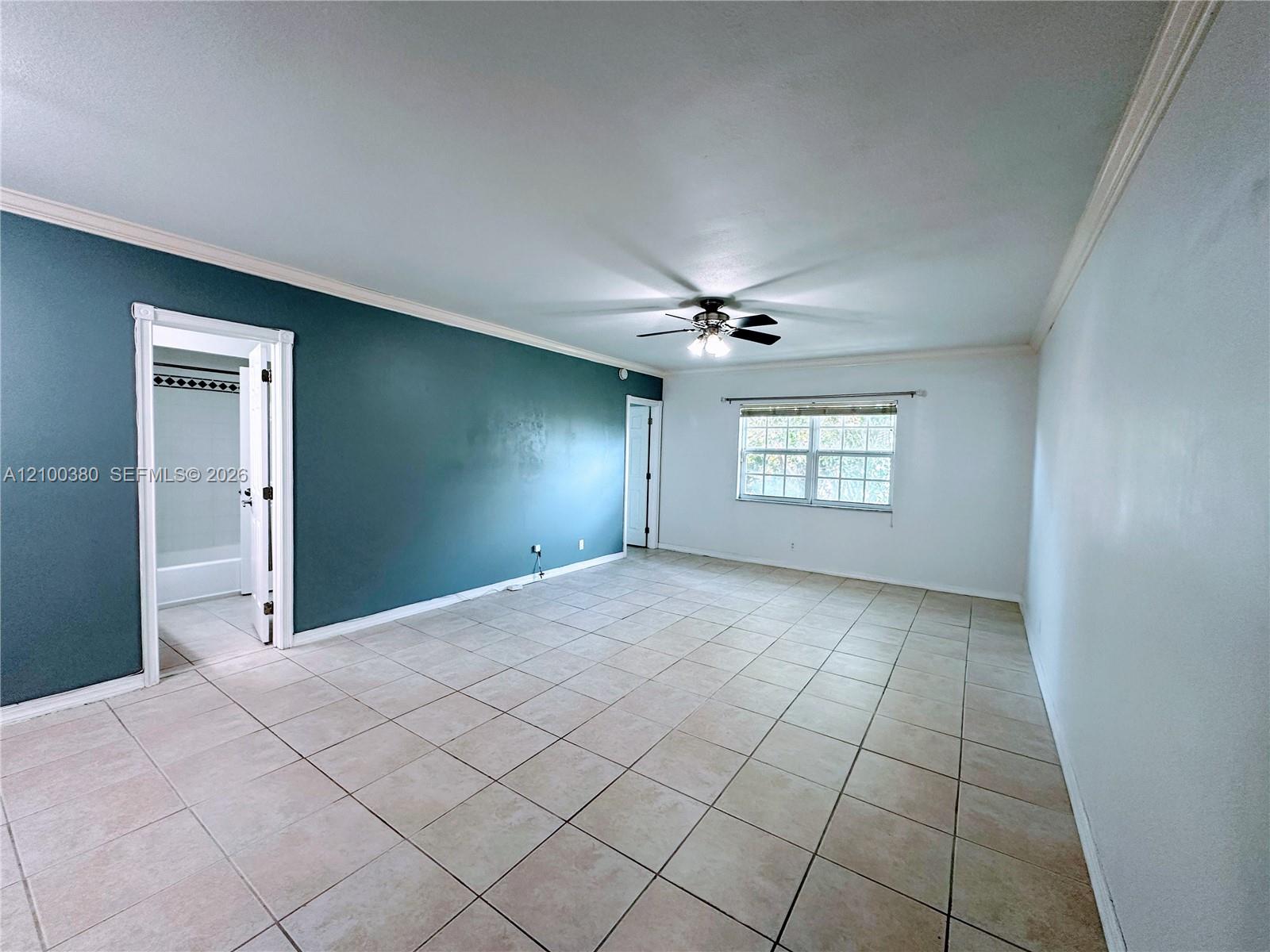 Empty tiled room with a teal accent wall, ceiling fan, window, and doorway to a bathroom.