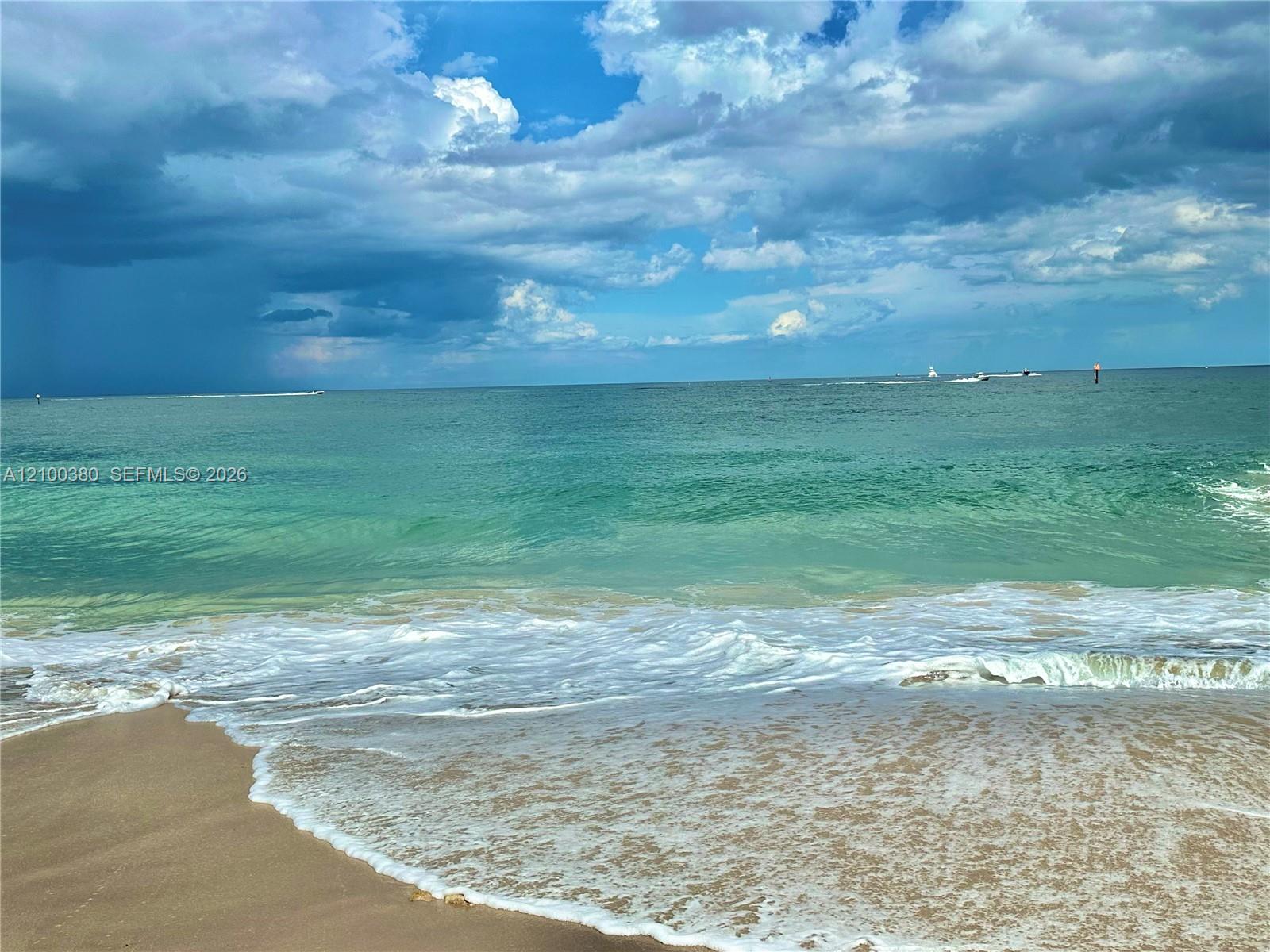Beach and turquoise ocean with distant boats beneath dramatic clouds.