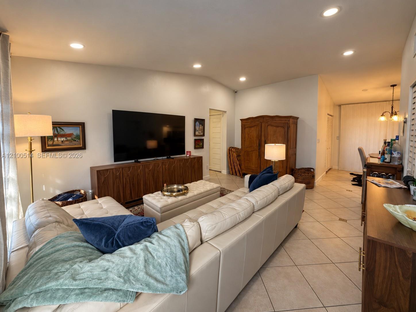 Living room with a white sectional, wall-mounted television, wooden console, and tiled floors.