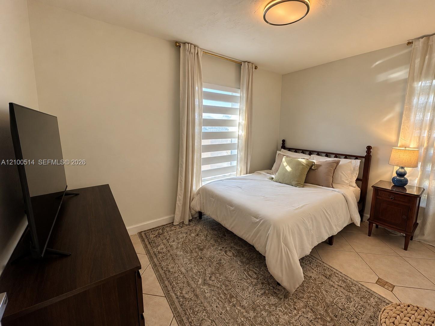 Bedroom with a white bed, patterned rug, dark wood furniture, and a curtained window.