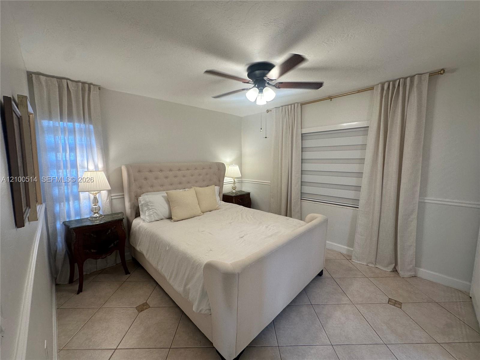 Bedroom with a cream upholstered bed, tiled floor, ceiling fan, and curtained windows.