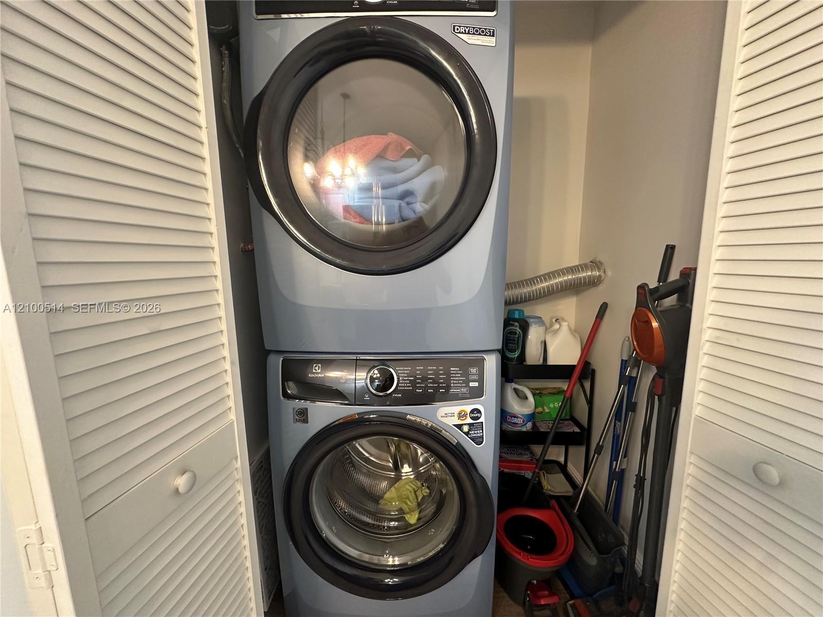 Stacked washer and dryer in a laundry closet, with cleaning supplies and tools stored alongside.