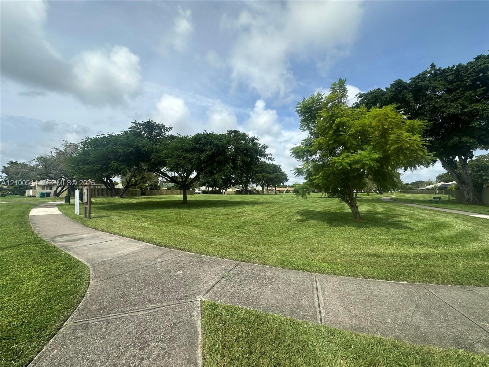 Community green space with a curving concrete path, broad lawns, mature trees, and a bench.