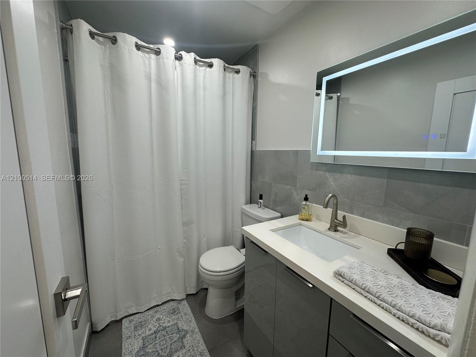 Bathroom with a gray vanity, illuminated mirror, and white shower curtain.