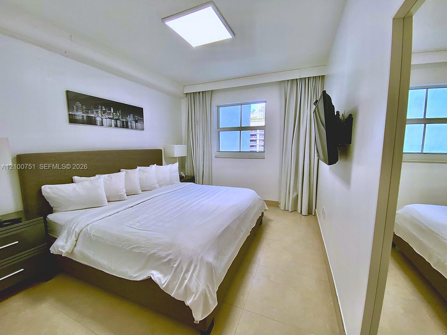 Bedroom with a large white bed, curtained window, and wall-mounted television.