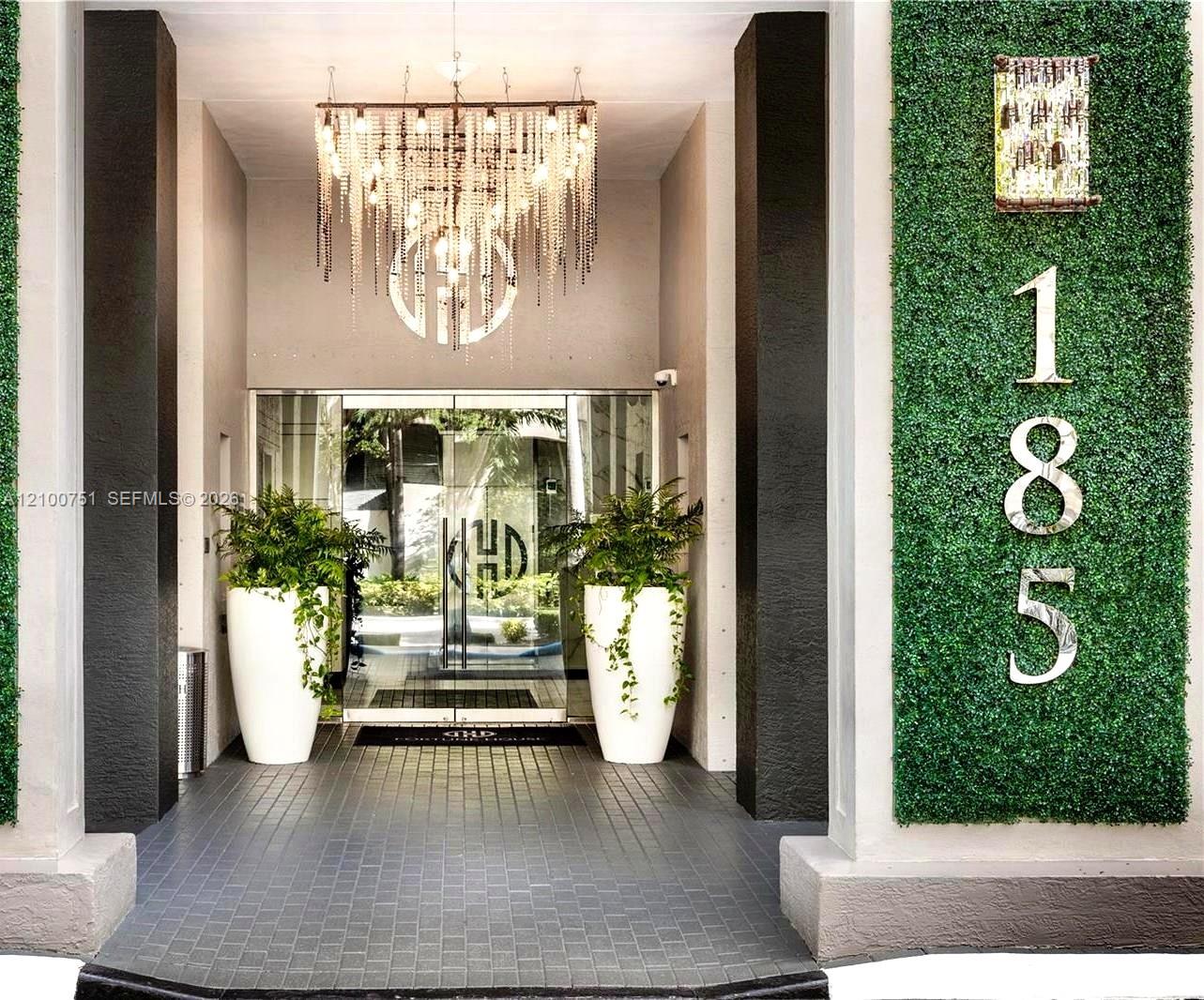 Modern building entrance with glass doors, hanging chandelier, potted plants, and a green wall marked 185.