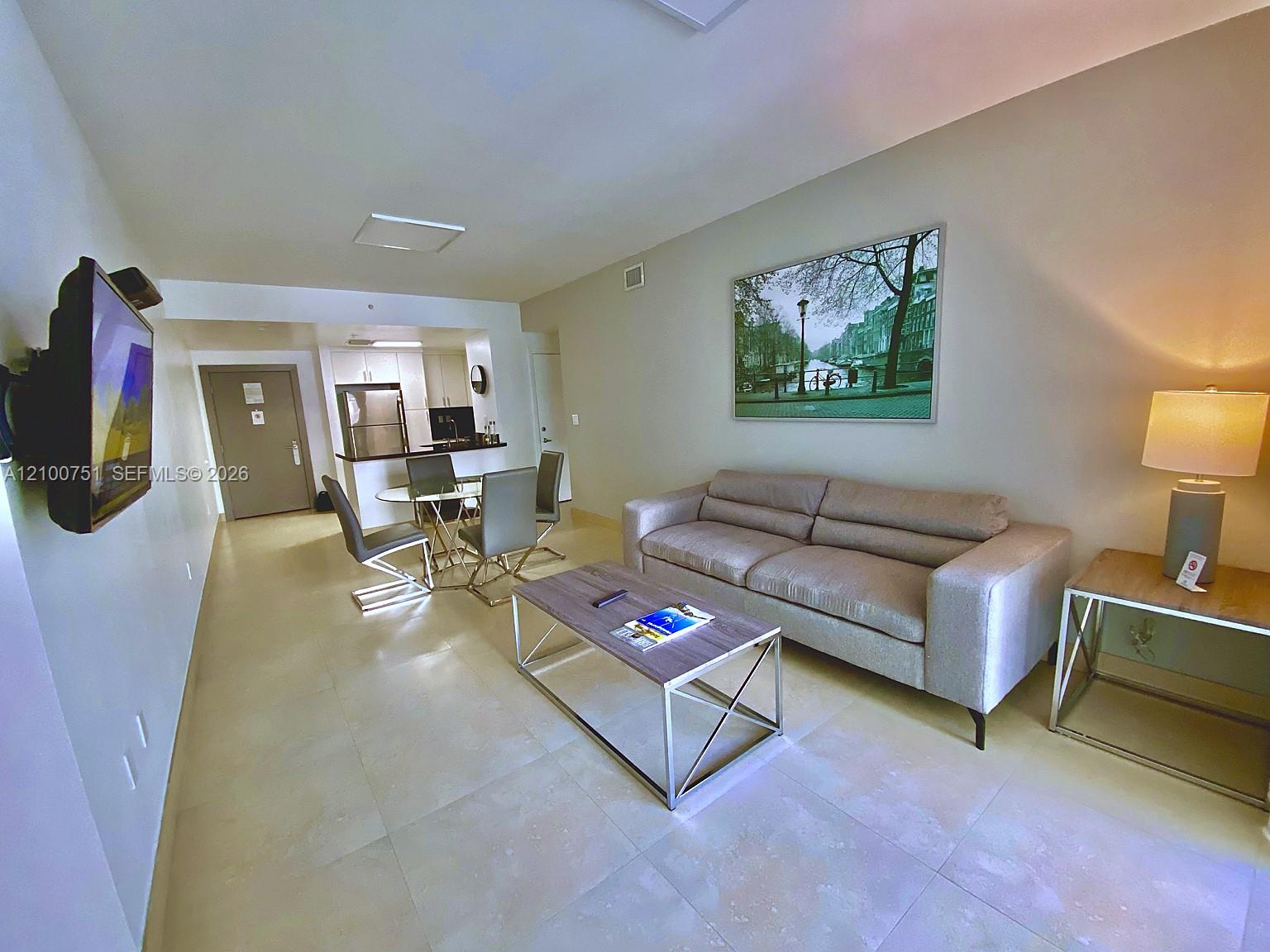 Furnished living room with a gray sectional sofa, glass dining table, and compact kitchen.