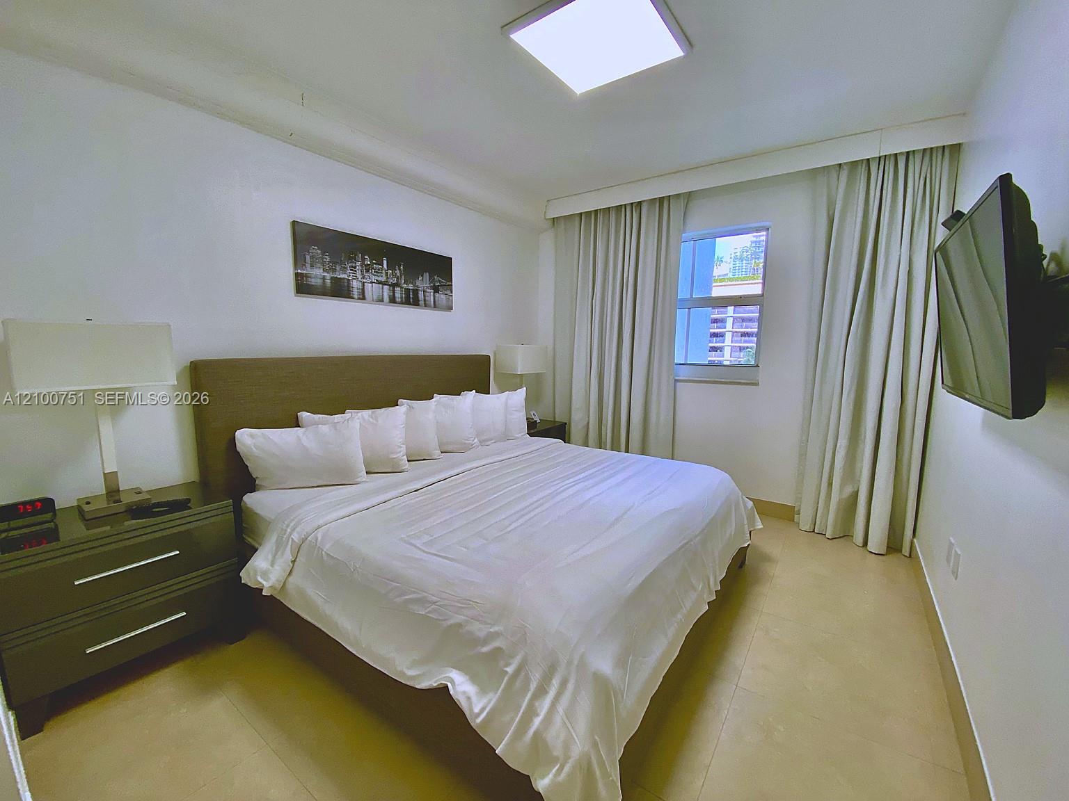 Furnished bedroom with a white bed, dark nightstands, floor-length curtains, and a wall-mounted TV.