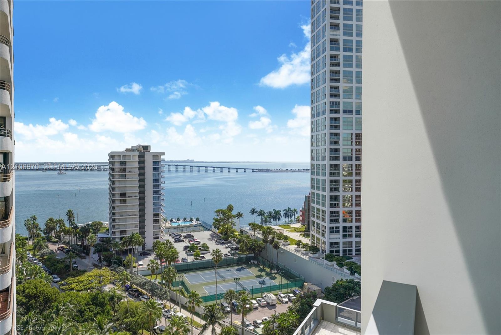 High-rise balcony view across Biscayne Bay, with a bridge, neighboring towers, and tennis courts below.
