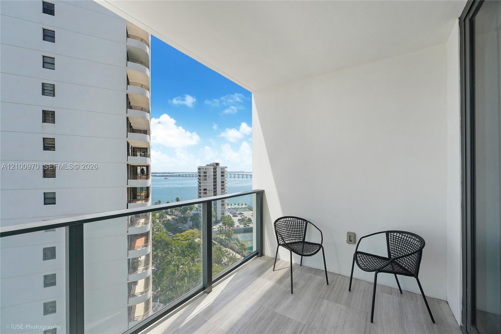 Apartment balcony with two black chairs and a glass railing overlooking Biscayne Bay and a bridge.