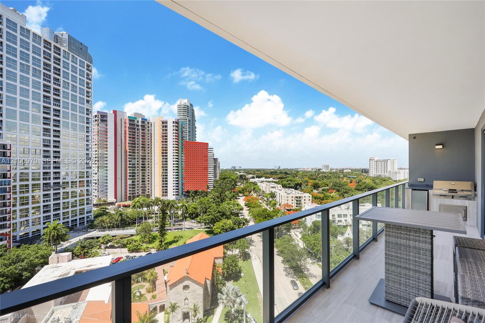 High-rise balcony with outdoor dining set and grill overlooking Miami towers and leafy streets.