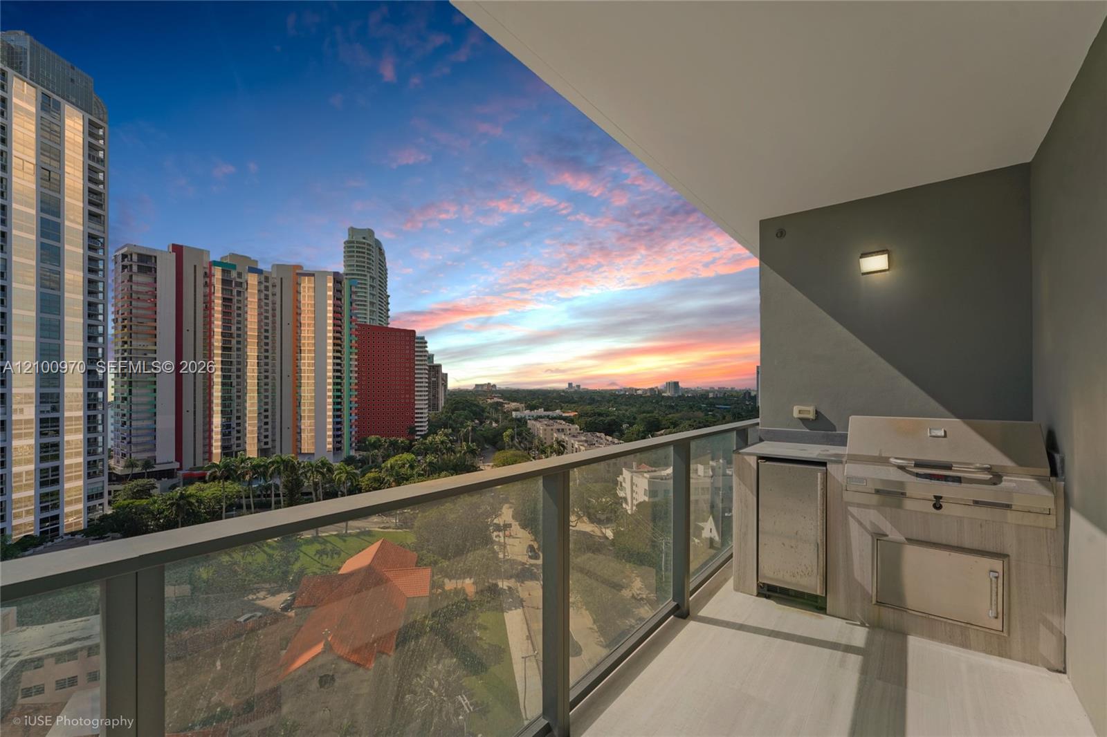 High-rise balcony with a built-in grill overlooking Brickell towers and greenery at sunset.