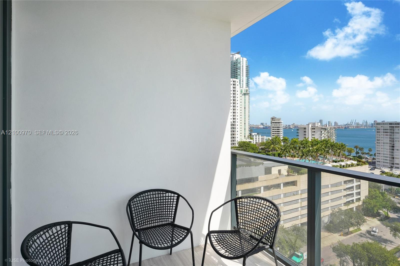 High-rise balcony with black woven chairs overlooking Biscayne Bay and Miami buildings.