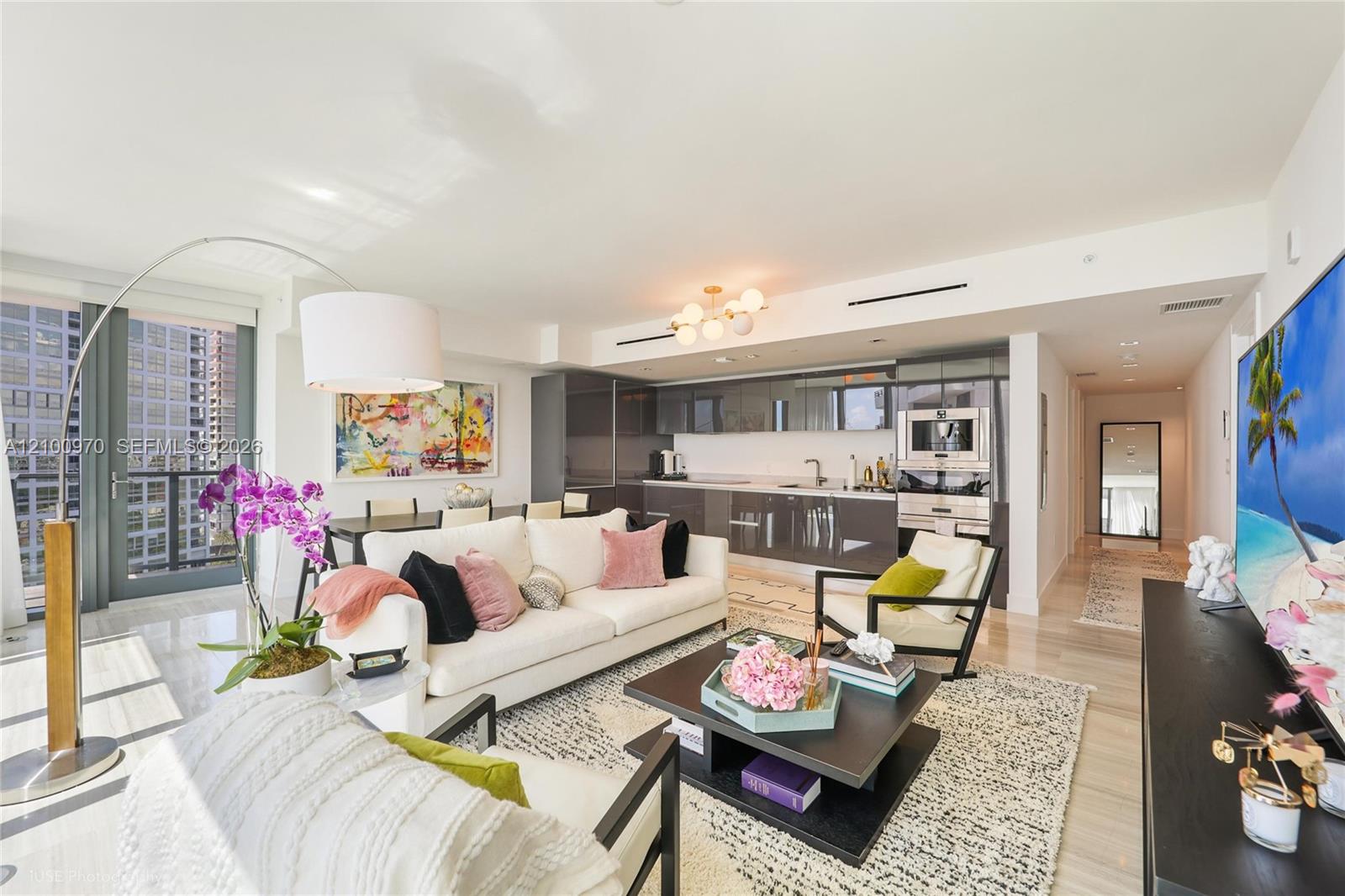 Furnished condo living room with white sofas, a dark modern kitchen, and floor-to-ceiling windows.