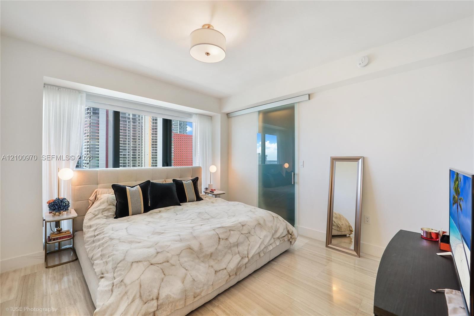 Modern condo bedroom with a marble-patterned bed and floor-to-ceiling windows overlooking high-rises.