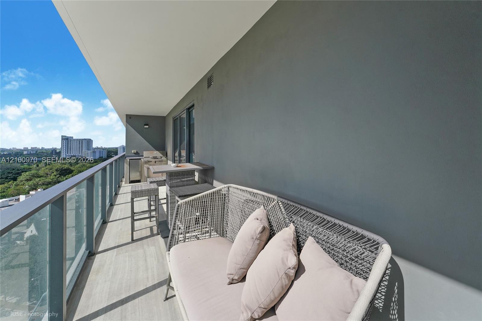Furnished condo balcony with woven lounge seating and views over green canopy and nearby buildings.