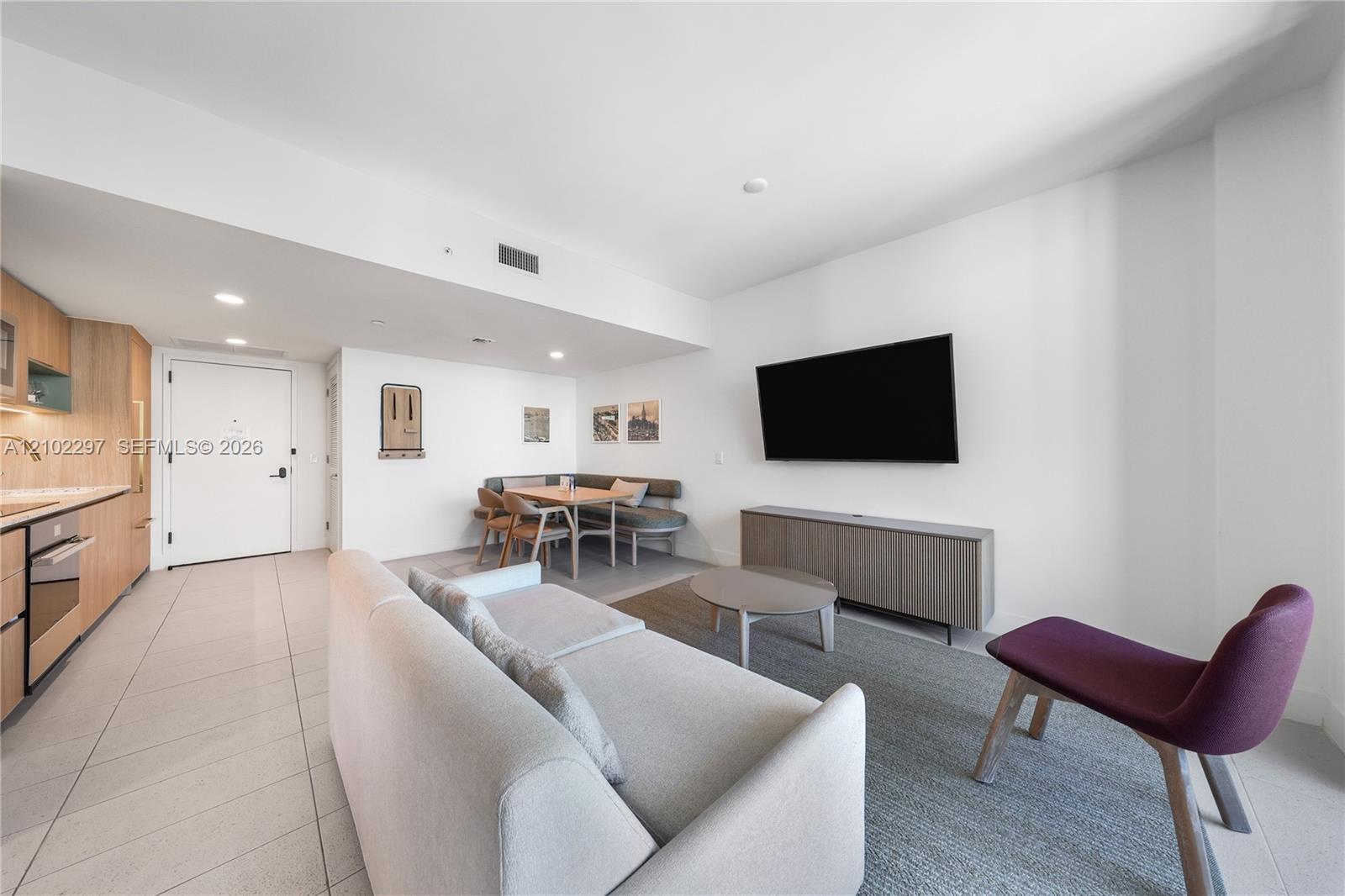 Furnished living room with a white sectional, wall-mounted TV, and dining nook beside the kitchen.