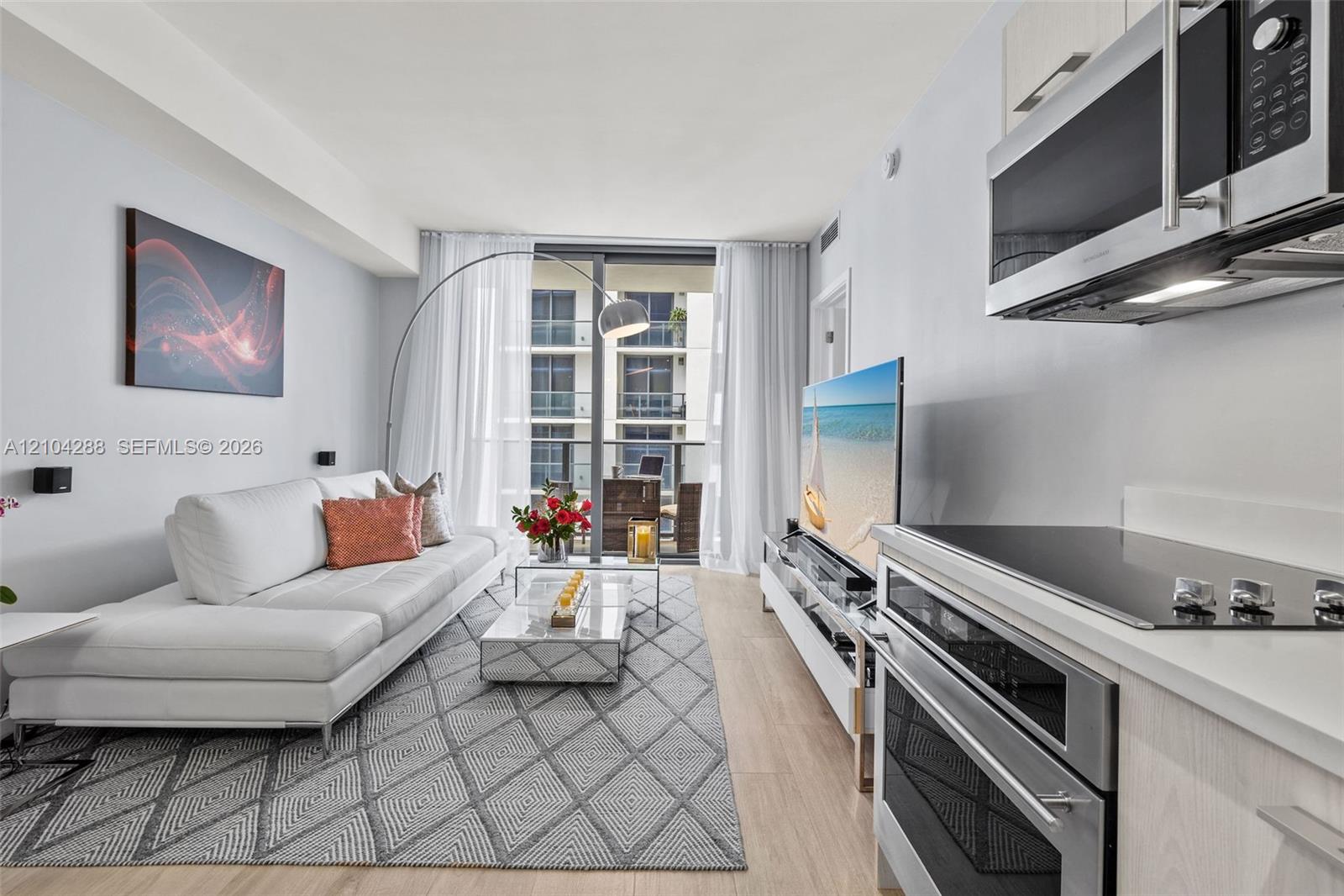 Furnished apartment living room with a white sectional, geometric rug, and stainless steel kitchen appliances.