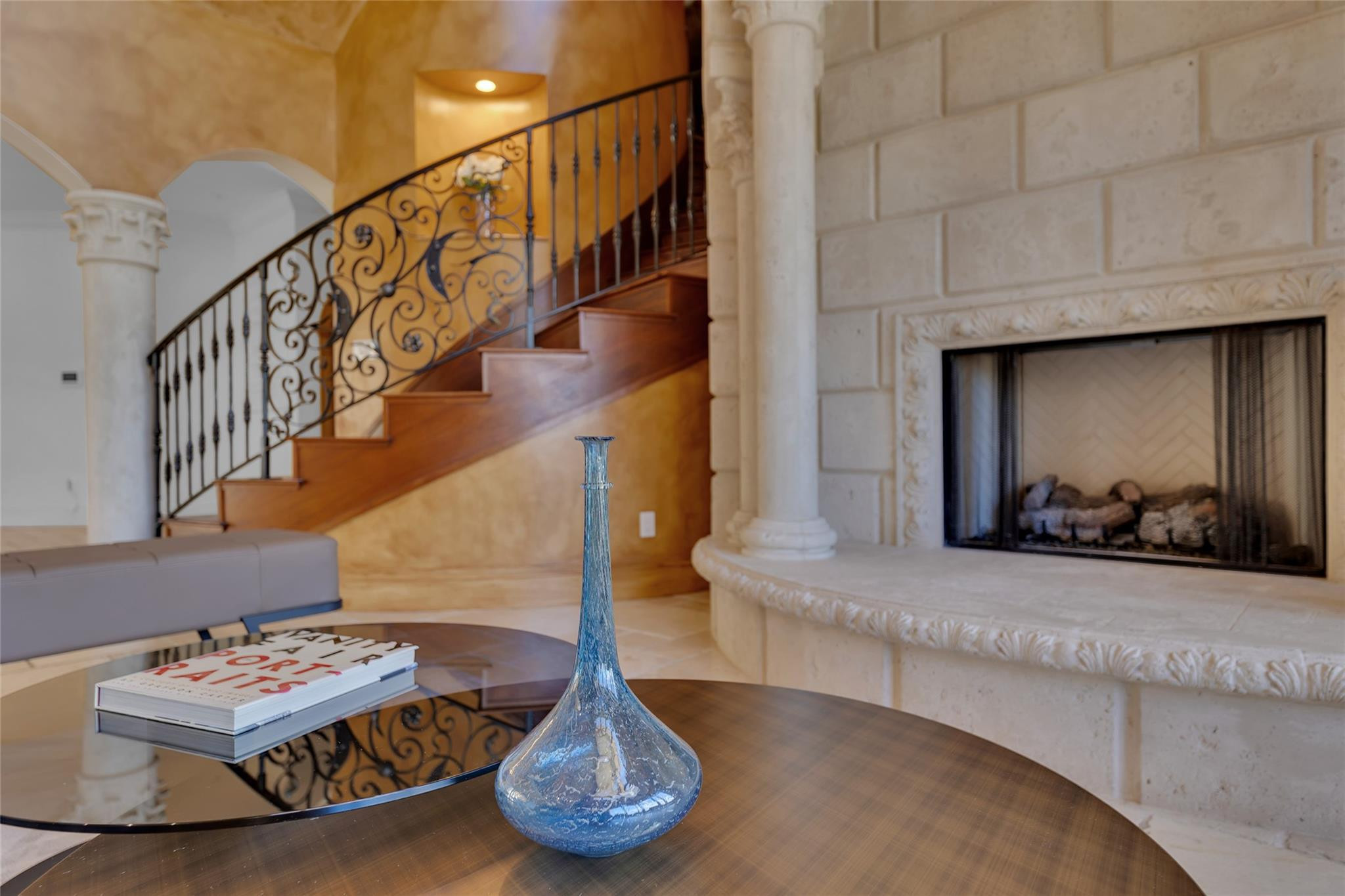 Living room with a carved stone fireplace, curved staircase, and glass coffee table.