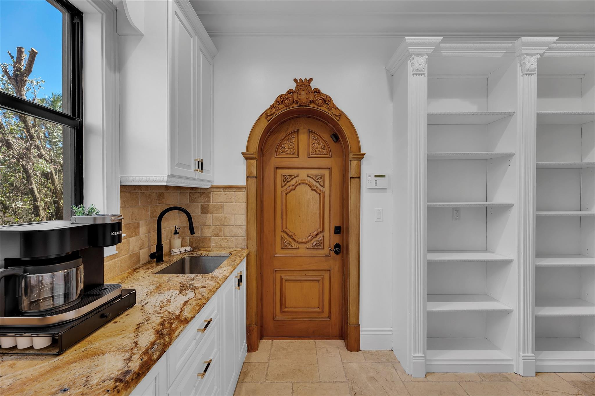 Kitchen coffee station with granite counters, built-in shelves, and an ornate arched wooden door.