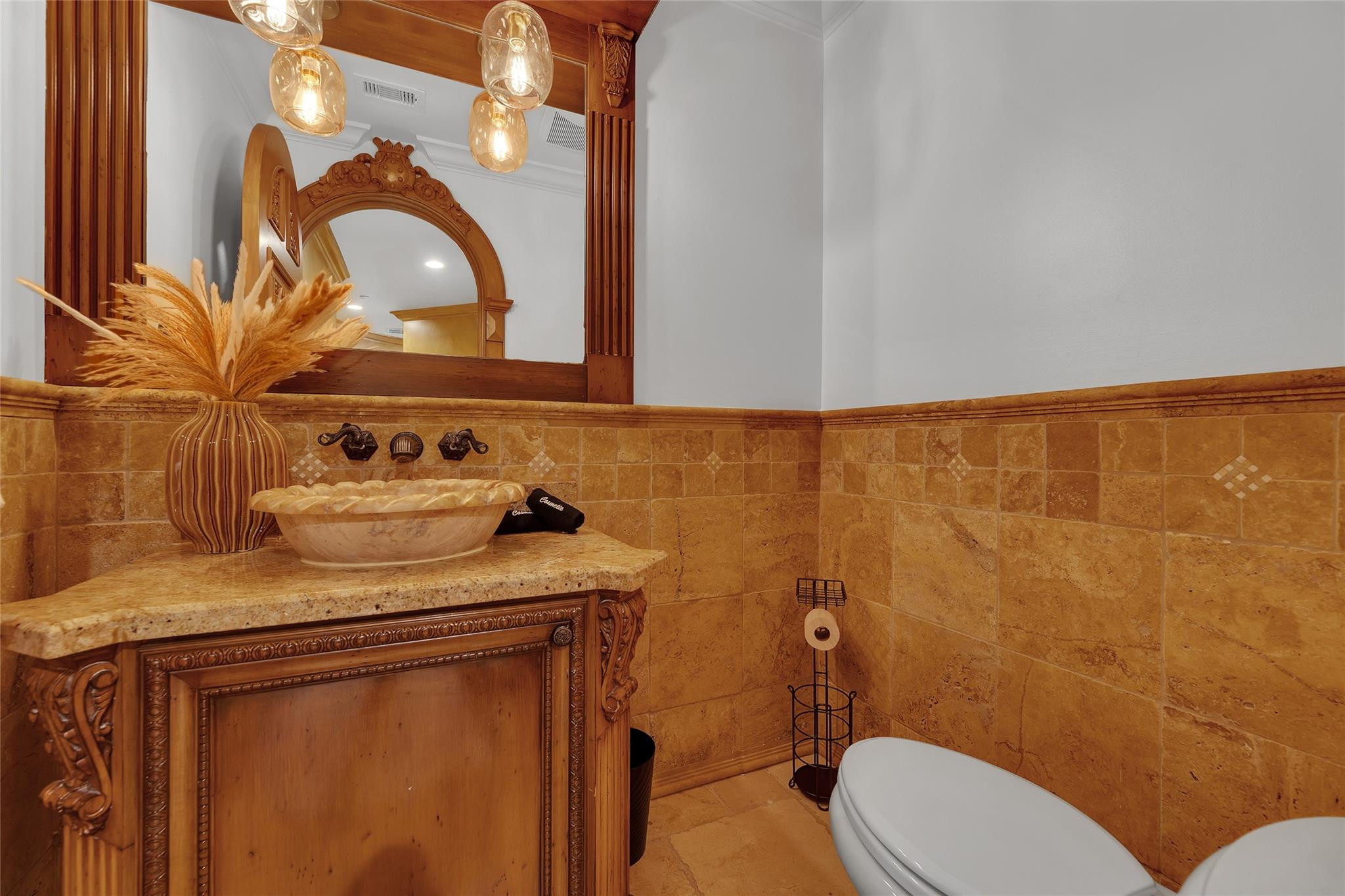 Bathroom with a carved wood vanity and vessel sink beside tan stone tile walls.