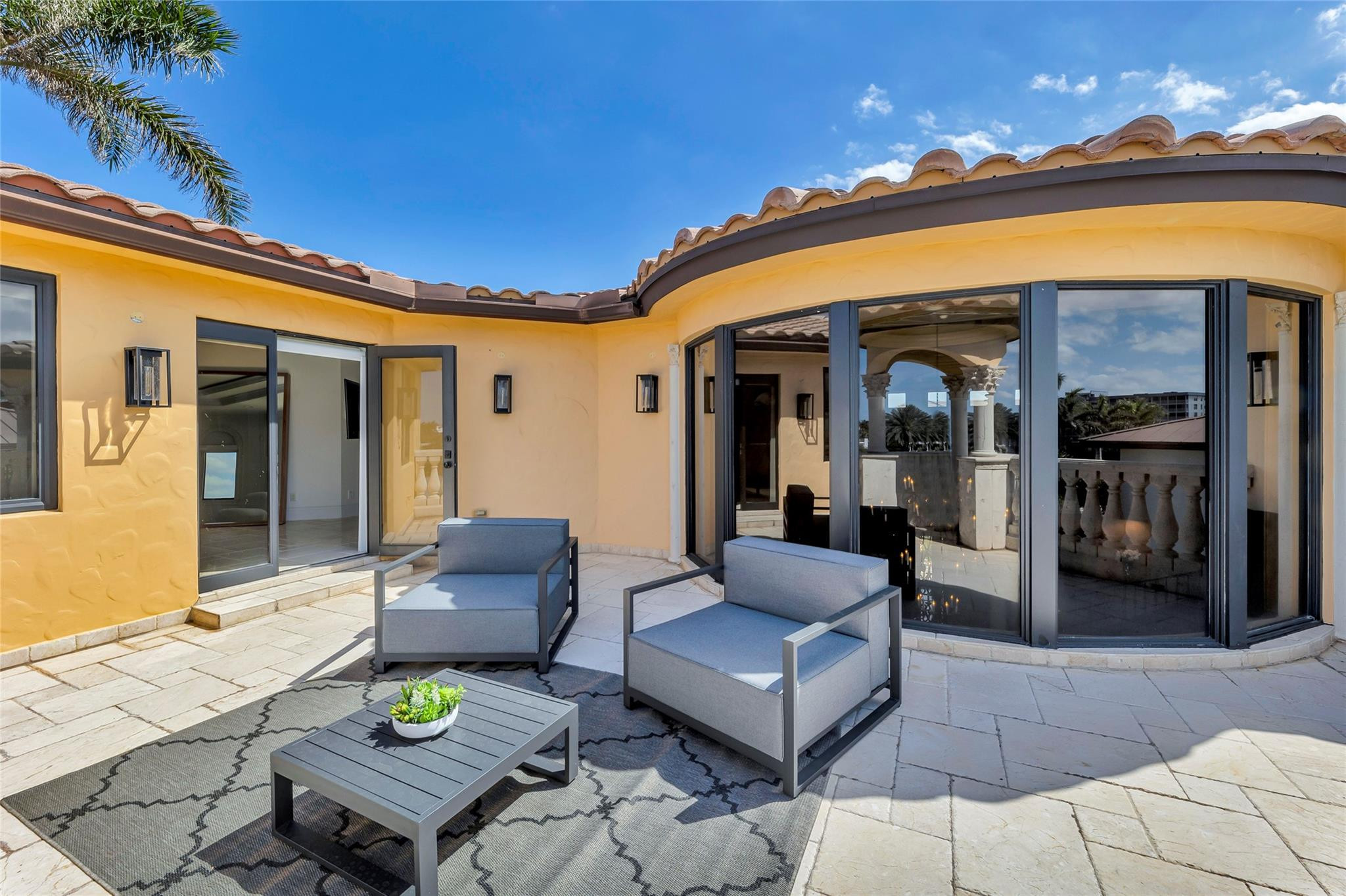 Stone patio with gray lounge seating beside curved glass doors and a terracotta tile roof.