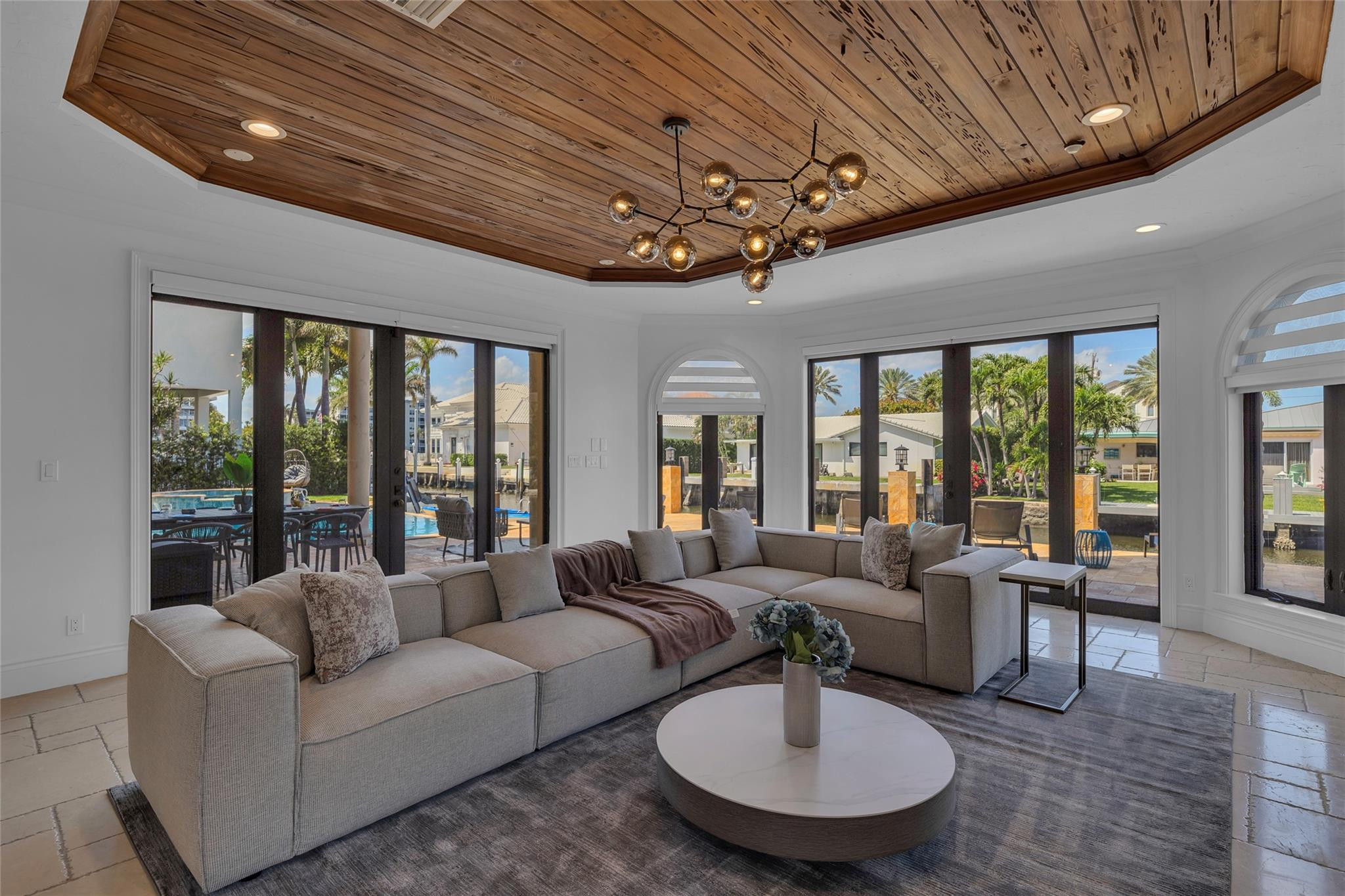 Living room with a wood-plank ceiling, sectional sofa, and glass doors overlooking a pool and canal.