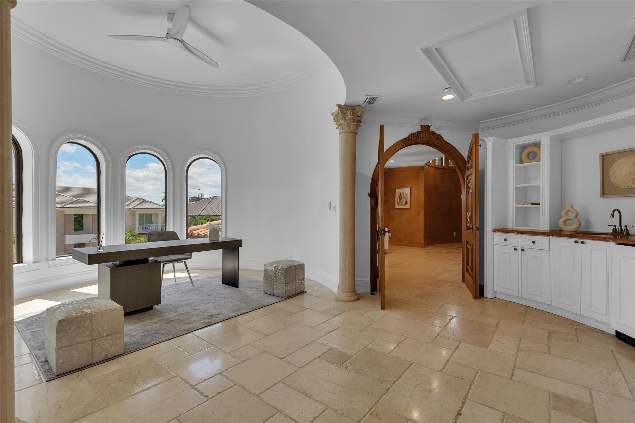 Home office with arched windows, a dark desk, stone floors, and a white wet bar.