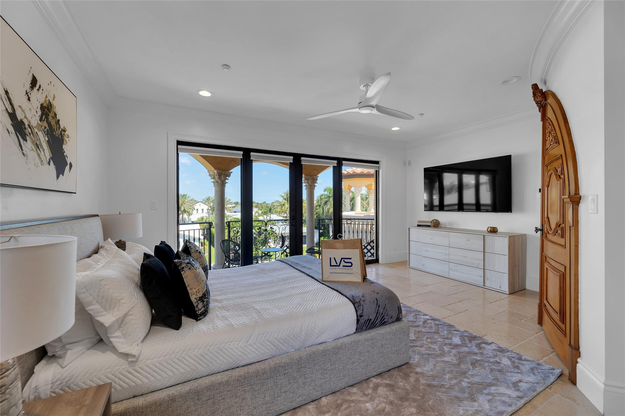Furnished bedroom with balcony doors, palm views, a wall-mounted television, and an ornate wooden door.
