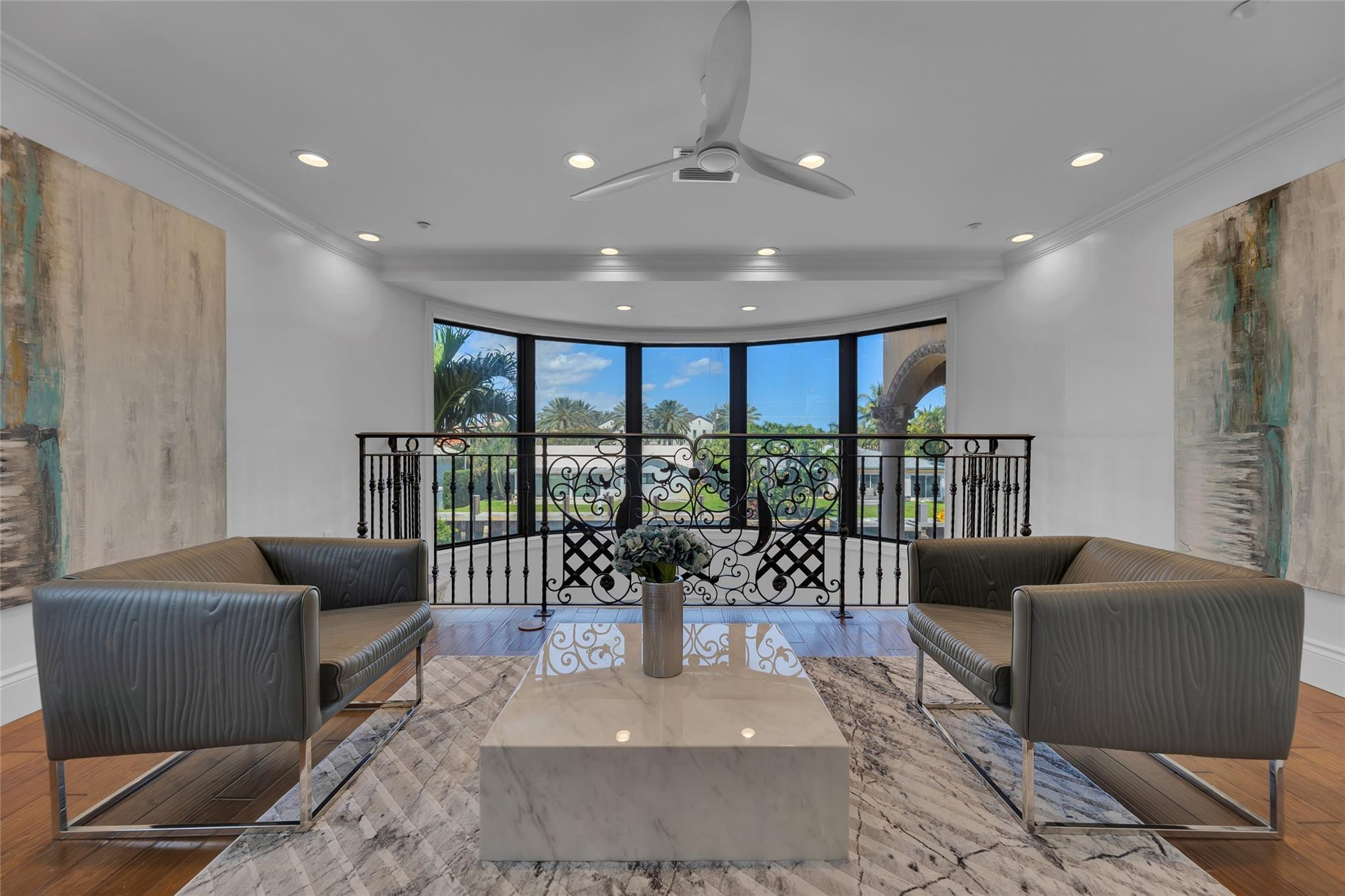 Furnished sitting area with gray armchairs and marble table beside tall windows overlooking a landscaped yard.