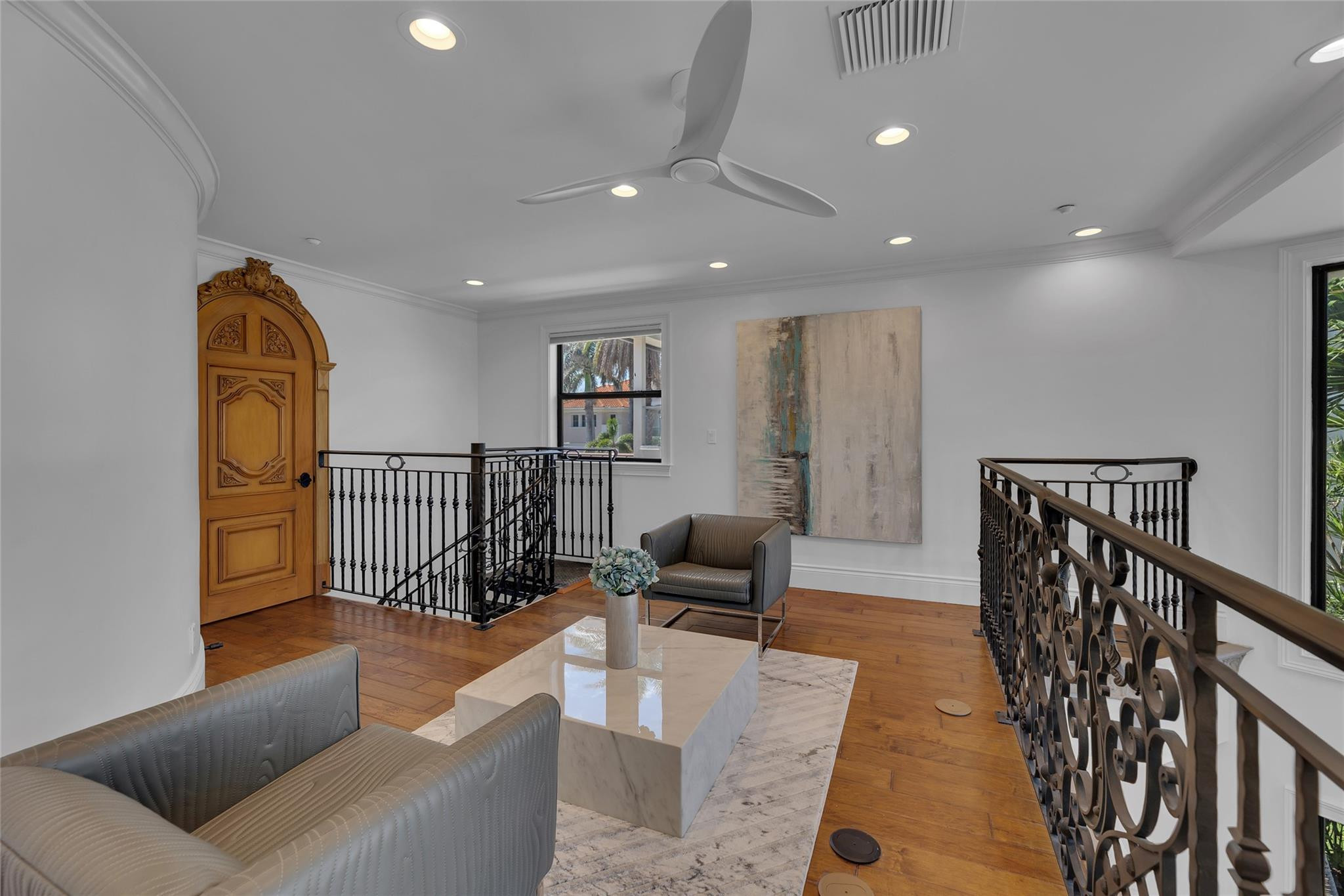 Upstairs sitting area with ornate iron railings, gray chairs, and a marble coffee table beside the stairwell.