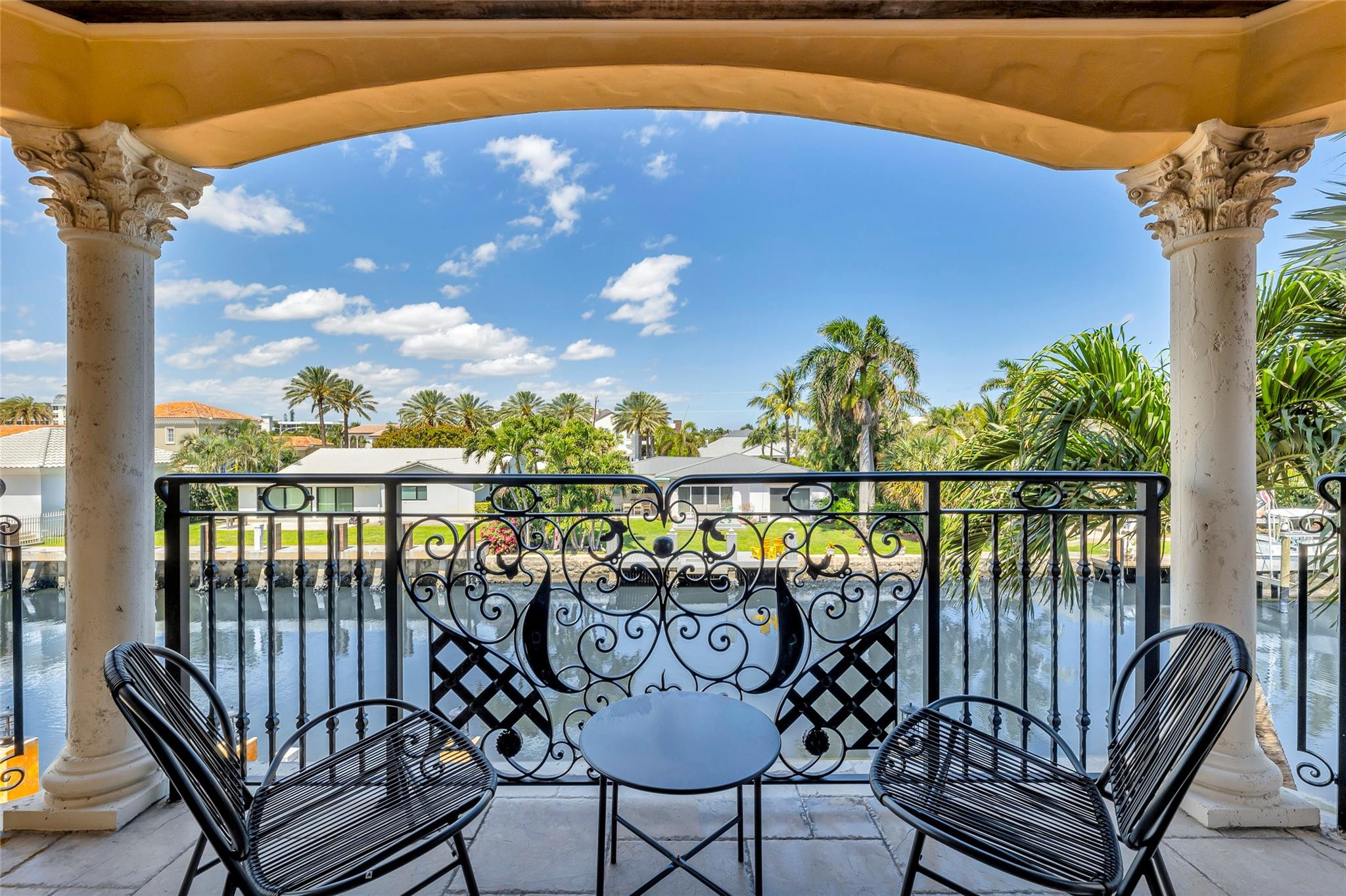 Covered balcony with ornate iron railing and seating overlooks a palm-lined canal.