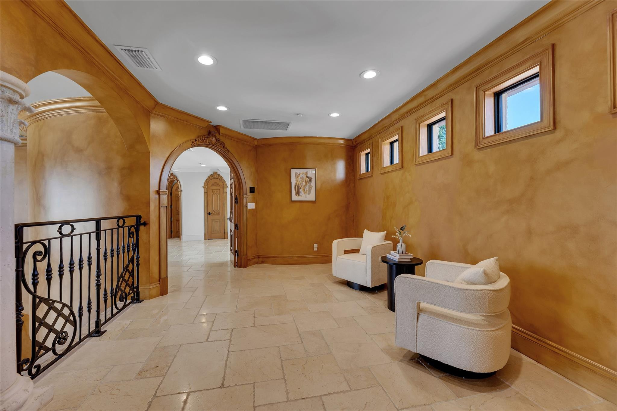 Foyer with ornate arched doorways, ochre walls, stone tile, and cream chairs beside high windows.