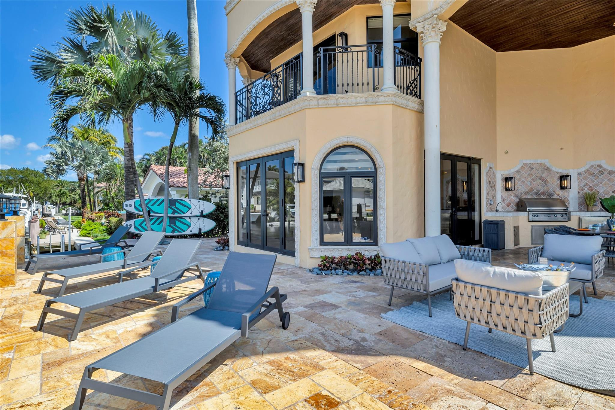 Furnished stone patio with an outdoor grill, palm trees, paddleboards, and an ornate balcony.