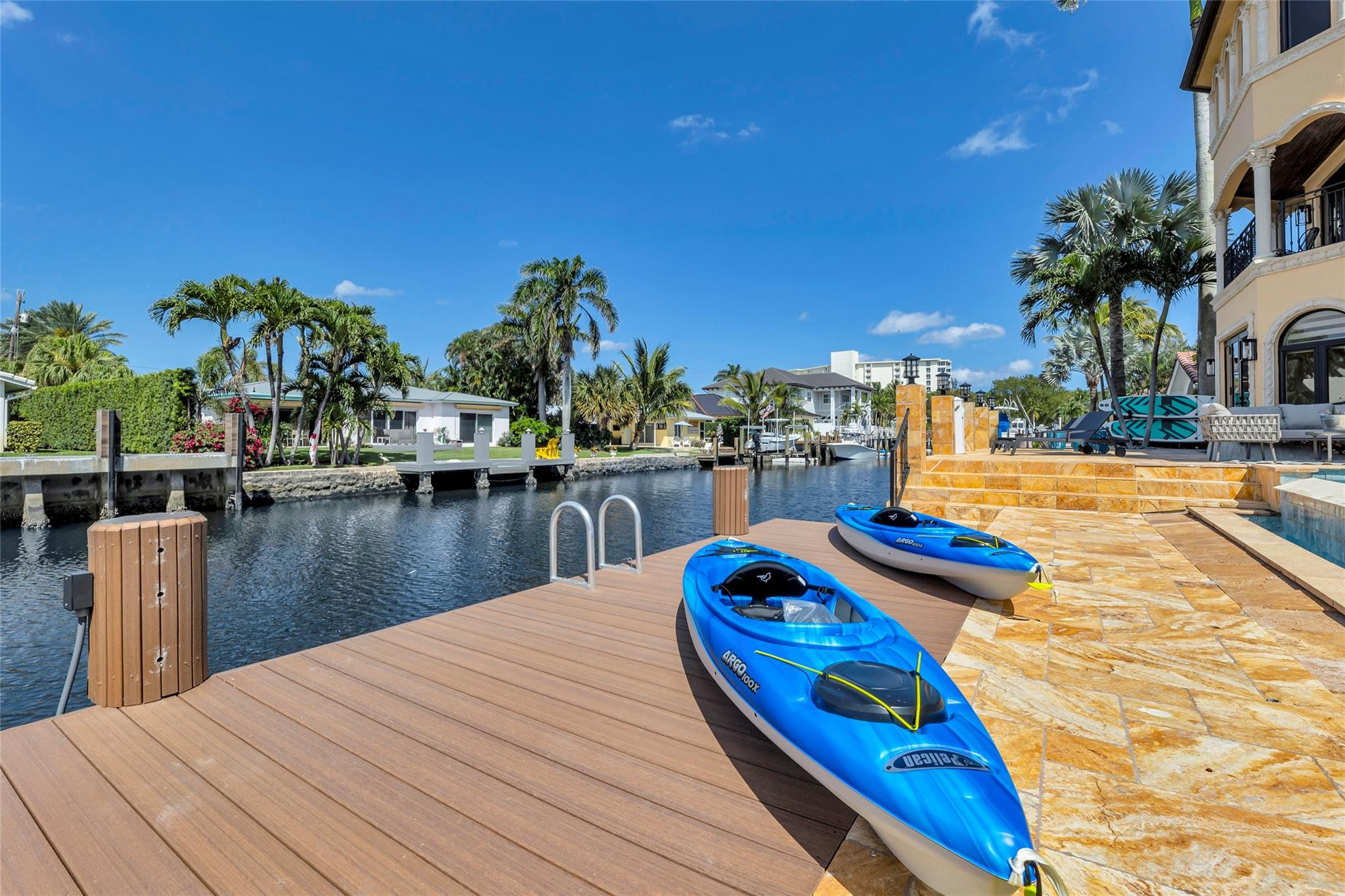 Waterfront dock and stone patio beside a canal, with two blue kayaks and palm trees.