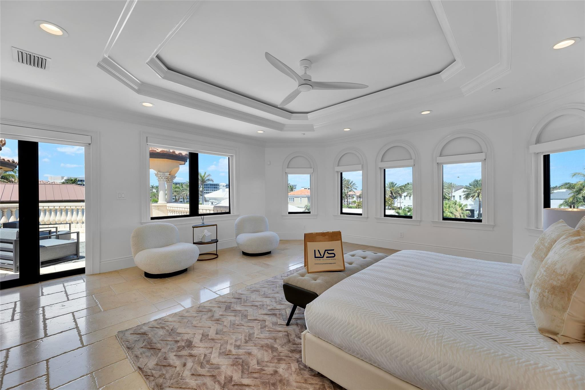 Bright bedroom with a white bed, arched windows, and views of palm trees and neighboring homes.
