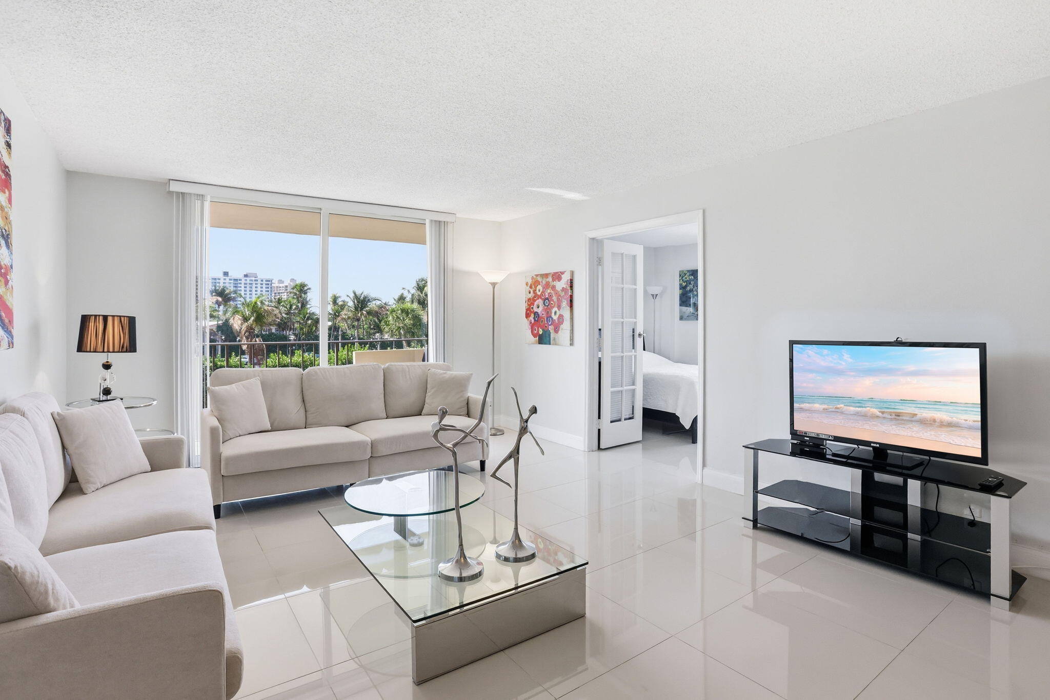 Furnished living room with large windows overlooking palm trees and nearby buildings.