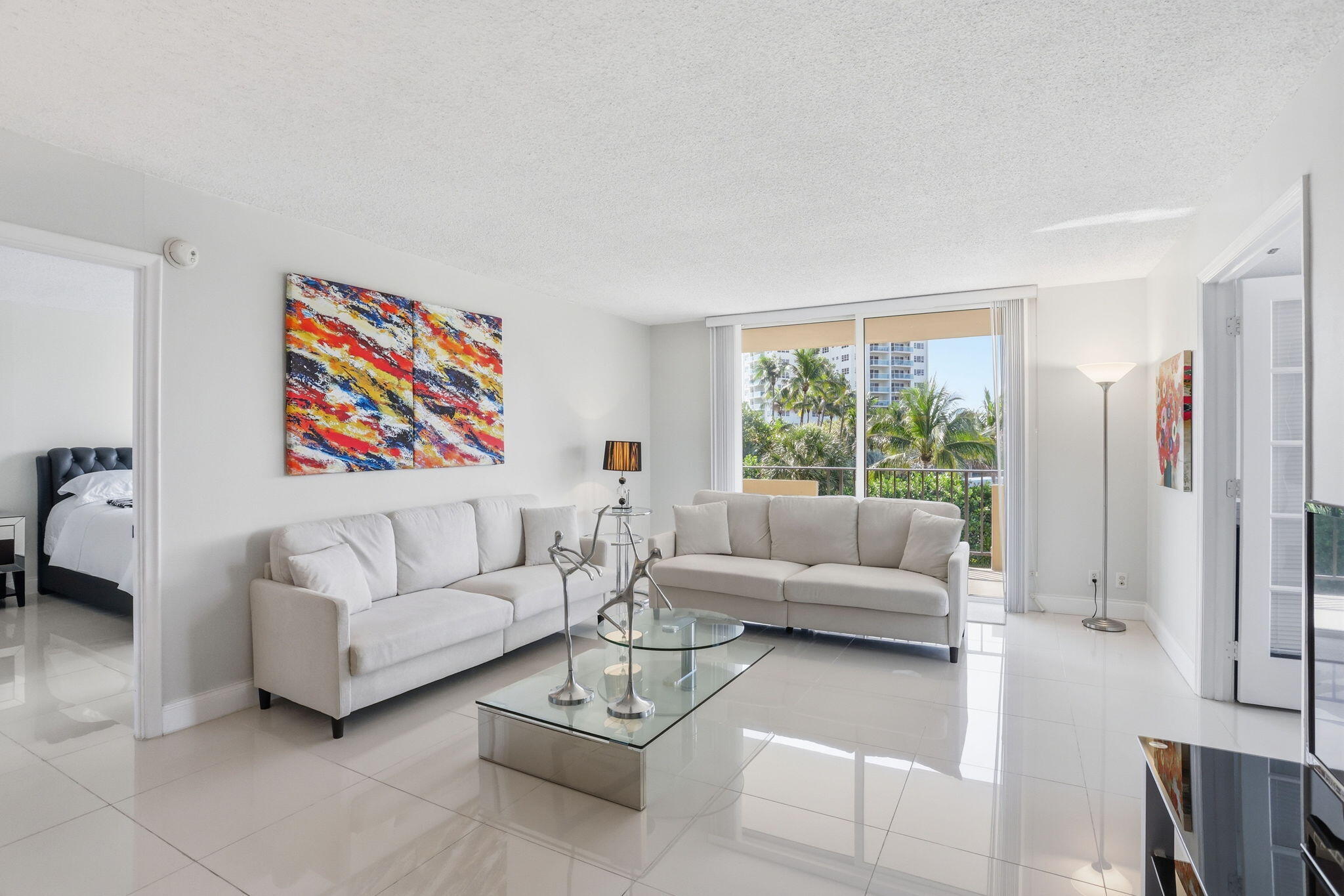 Bright condo living room with white sofas, colorful abstract art, and sliding doors overlooking palm trees.