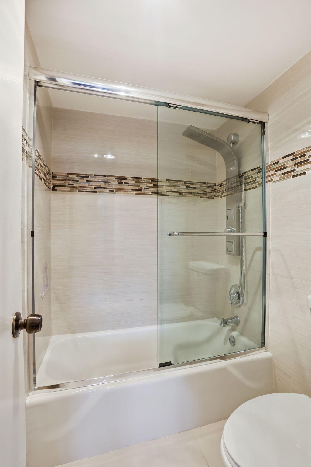Bathroom bathtub with glass shower doors, overhead showerhead, and mosaic tile accent band.