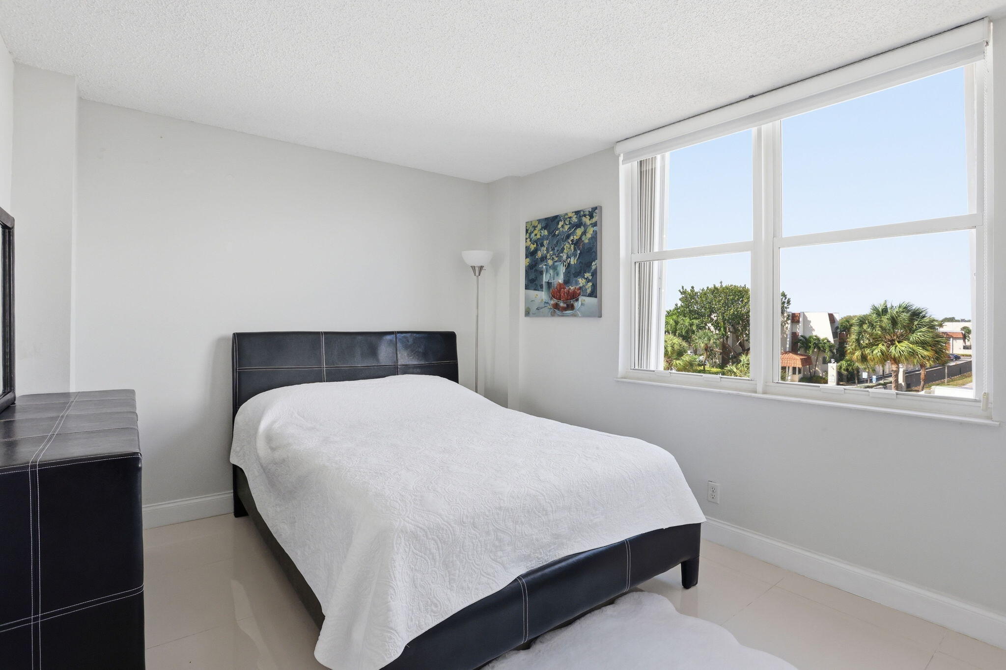 Bedroom with a black upholstered bed and large windows overlooking palm trees and nearby buildings.