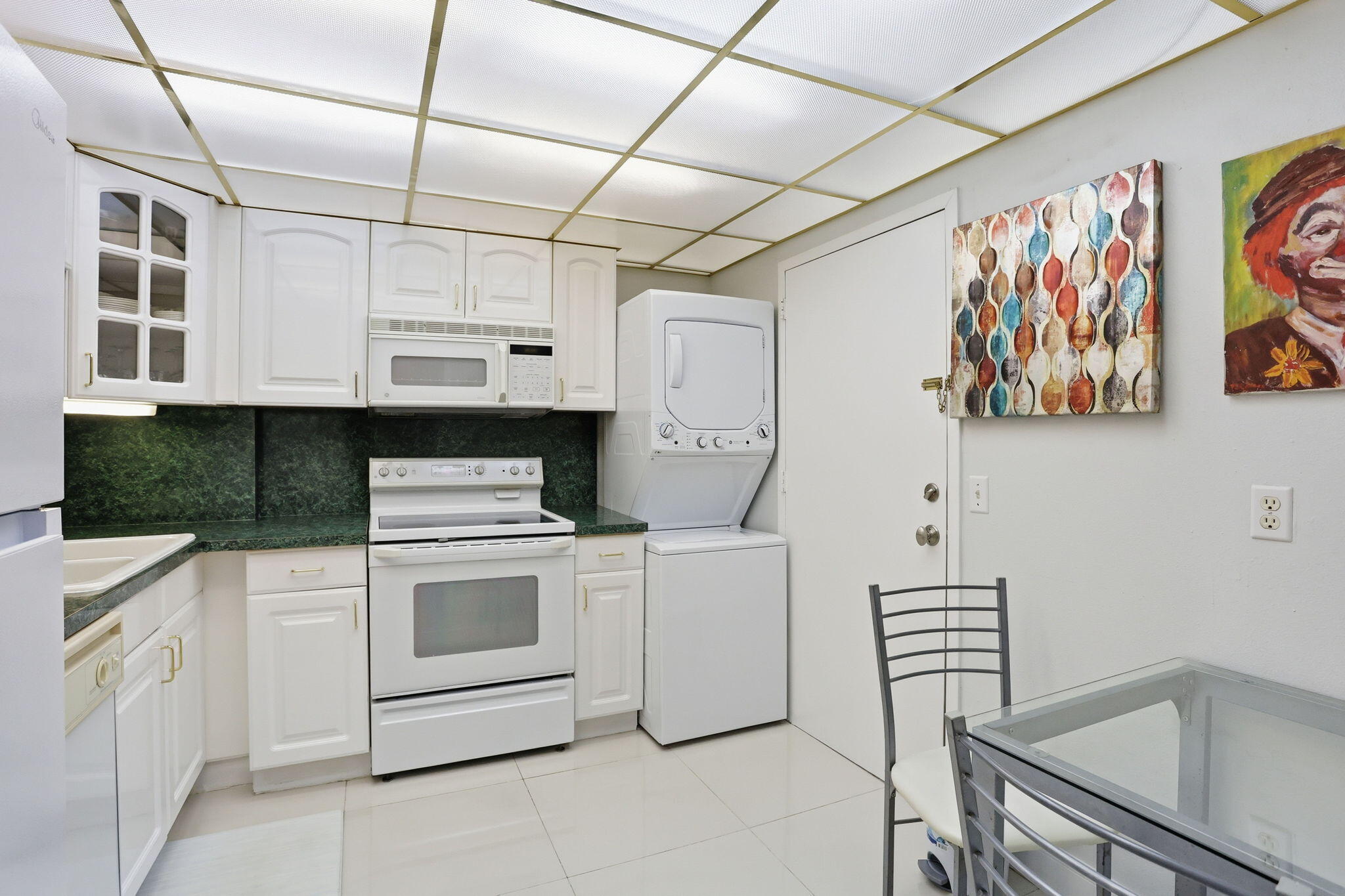 Kitchen with white cabinetry, dark green counters, stacked washer and dryer, and a glass-top dining table.