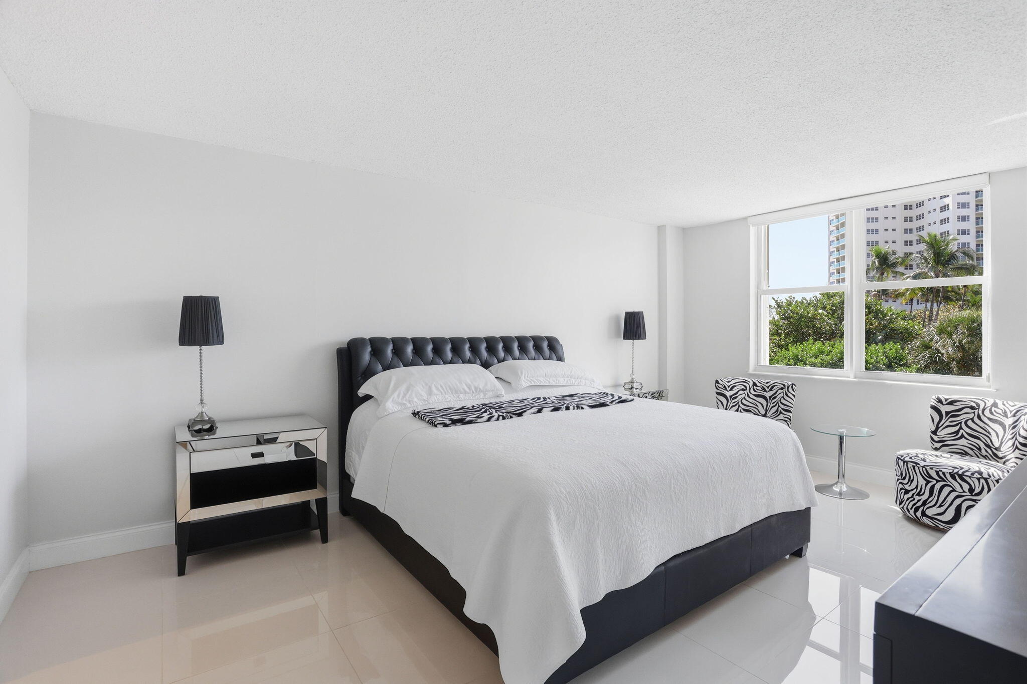 Bright bedroom with a black tufted bed, zebra-patterned chairs, and windows overlooking palm trees.