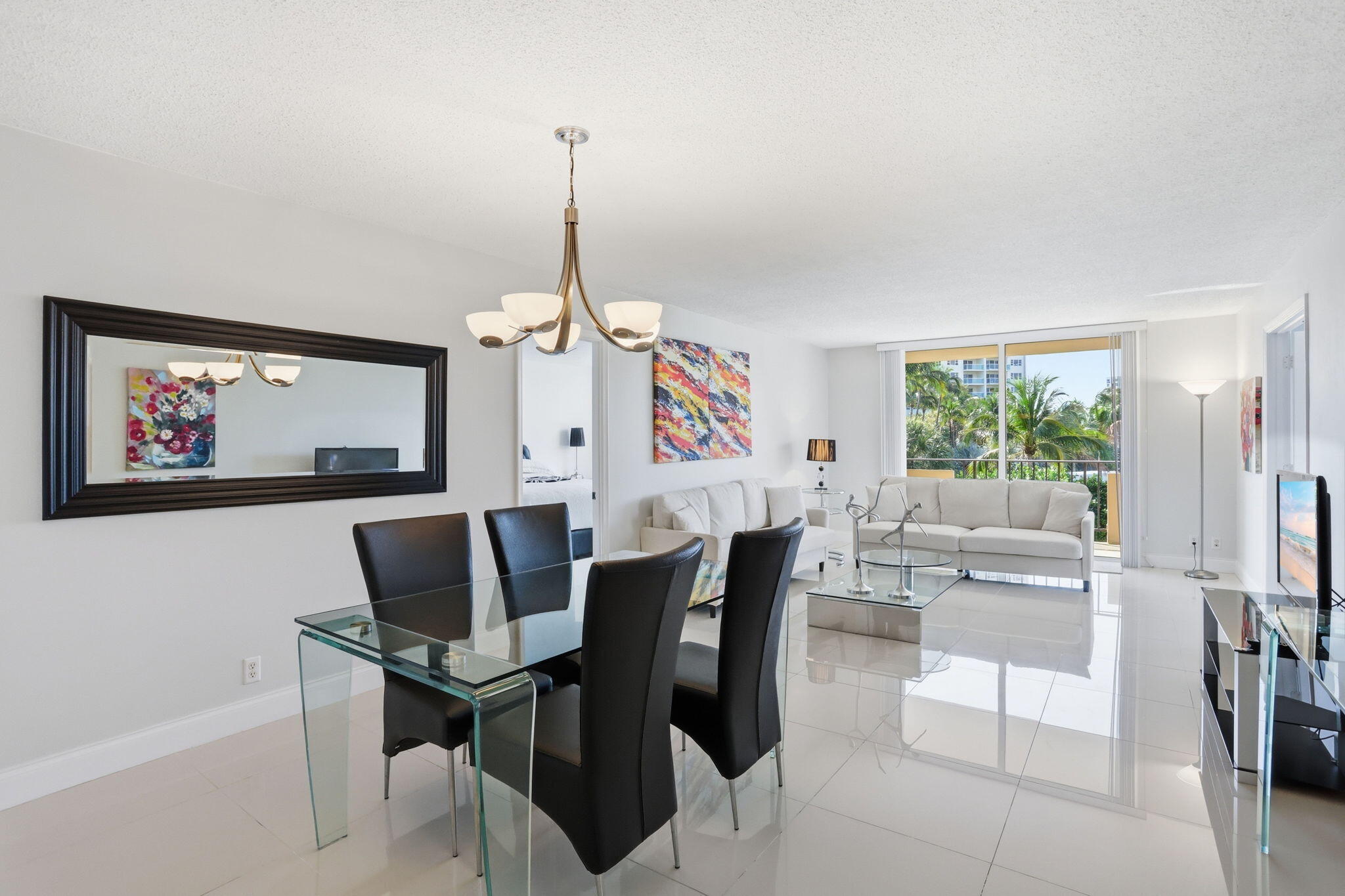 Open-plan living and dining room with white sofas, glass table, glossy tile floors, and palm views.